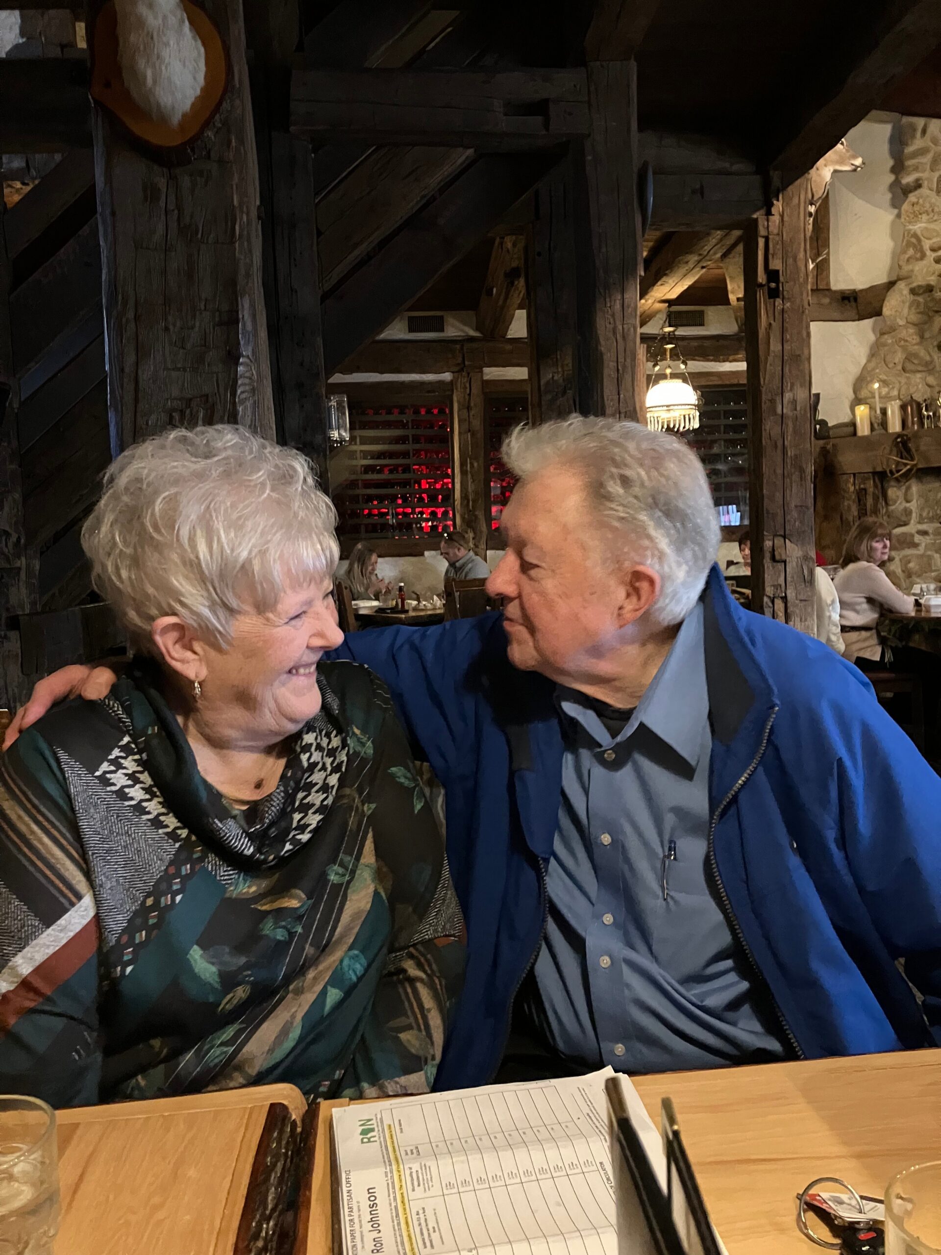 Two elderly people smiling and embracing at a rustic restaurant with dark wooden beams in the background.
