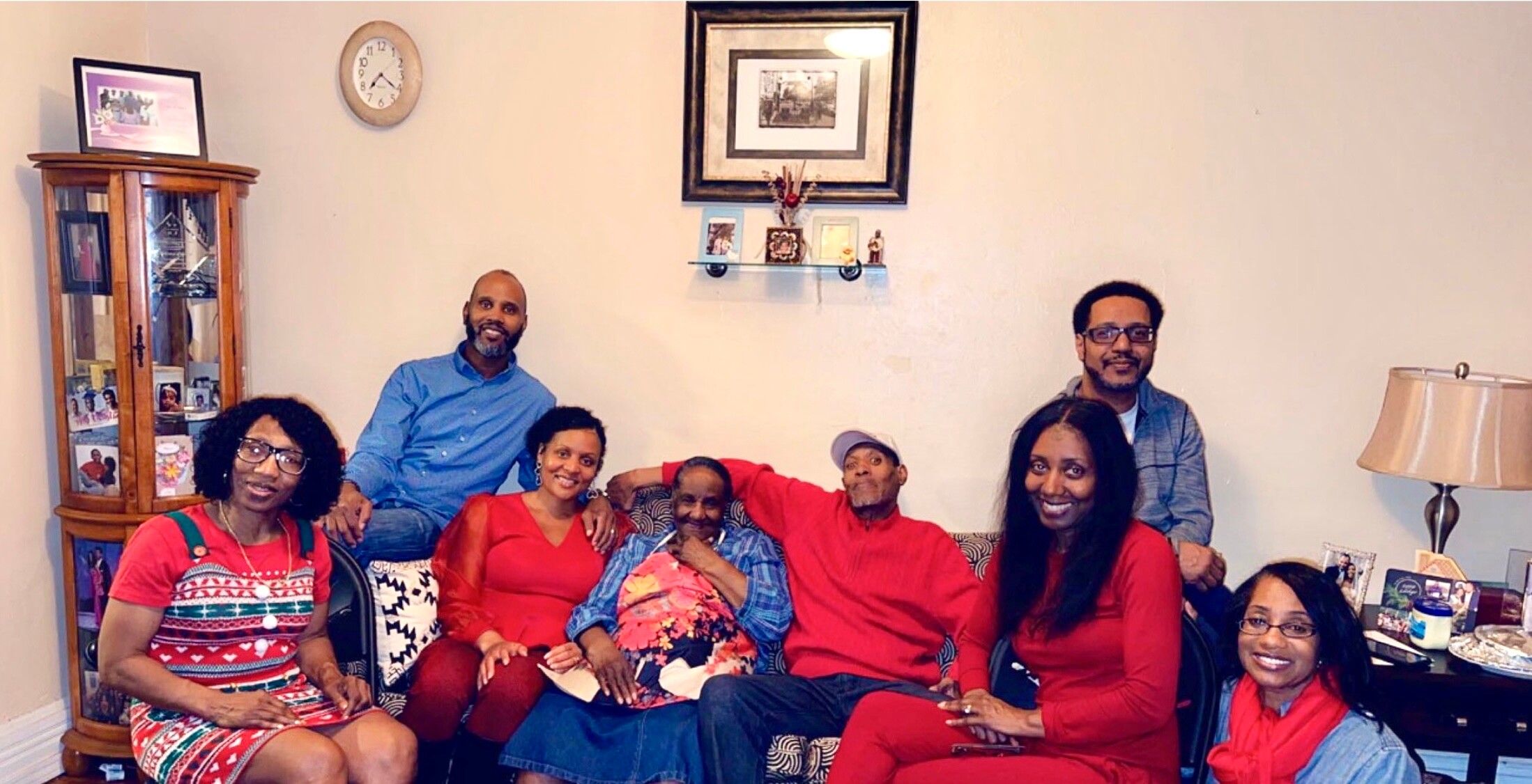 Group of eight adults posing together on a sofa in a living room, smiling at the camera, casual family photo in a home setting.
