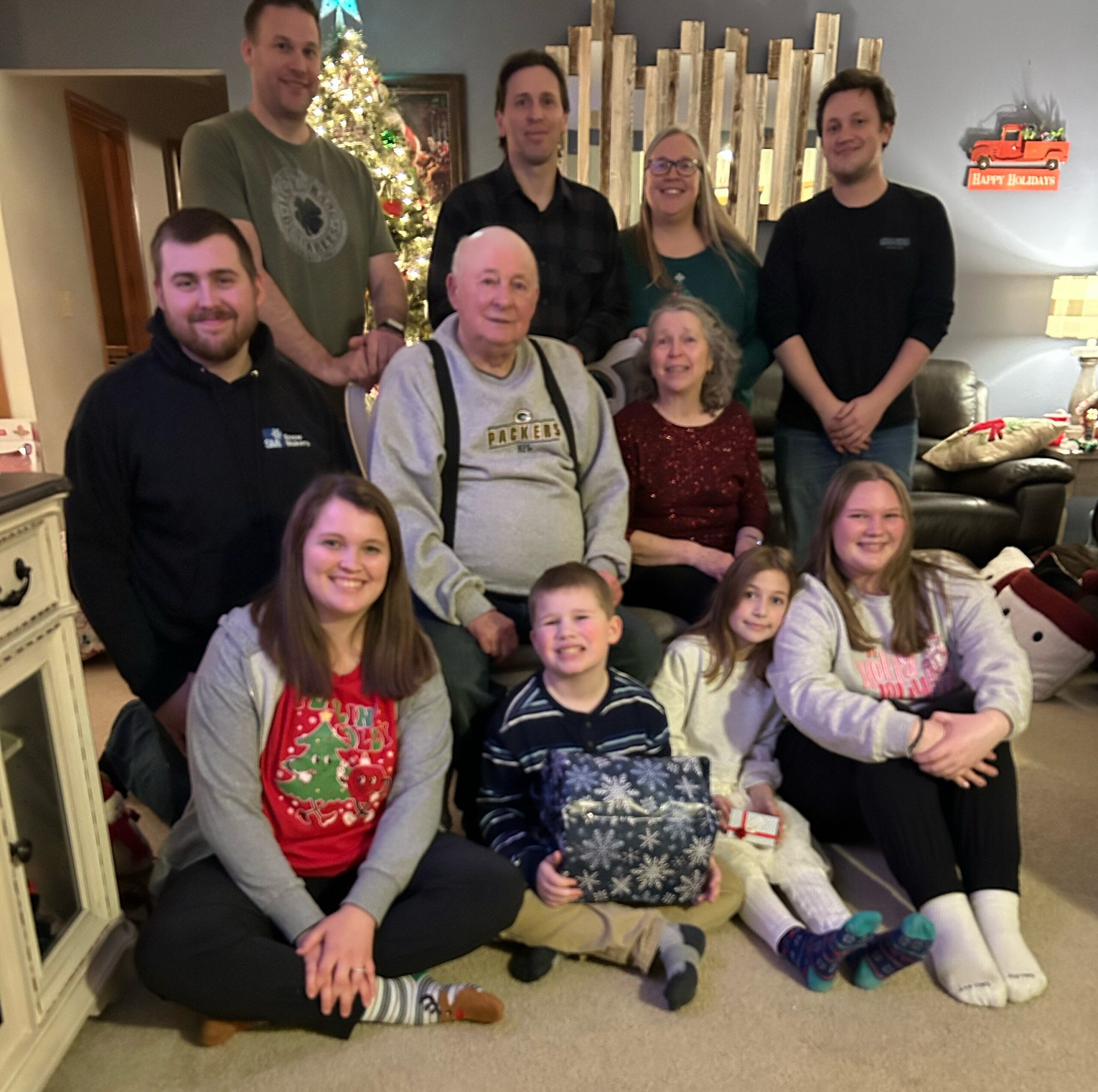 Group of family and friends posing for a Christmas photo in a living room, with a decorated tree in the background and wrapped gifts nearby.