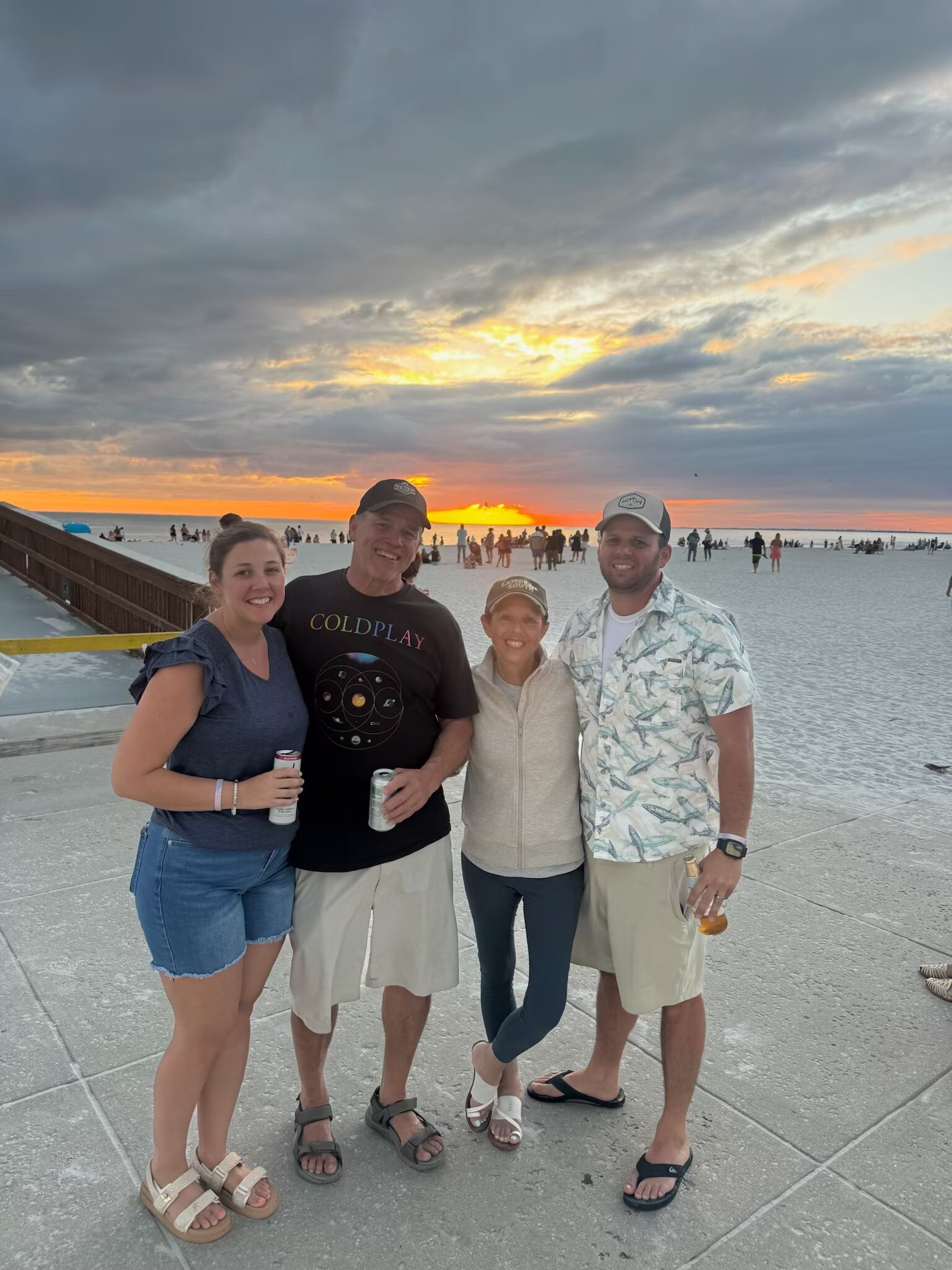 Four adults posing together on a beach boardwalk at sunset, holding drinks and smiling at the camera.