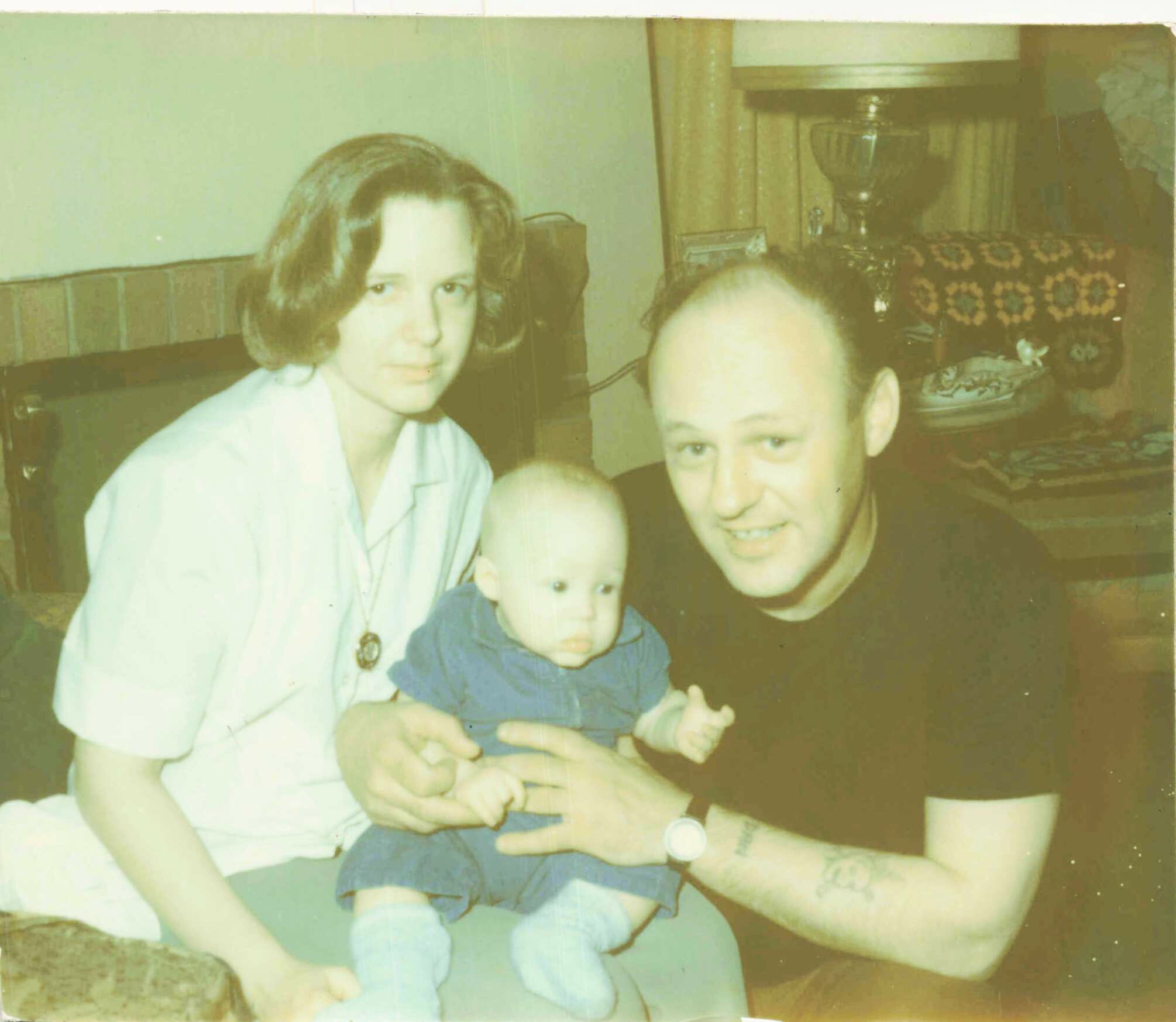 Family photo: a woman on the left holding a baby, with a smiling man beside them in a living room background.