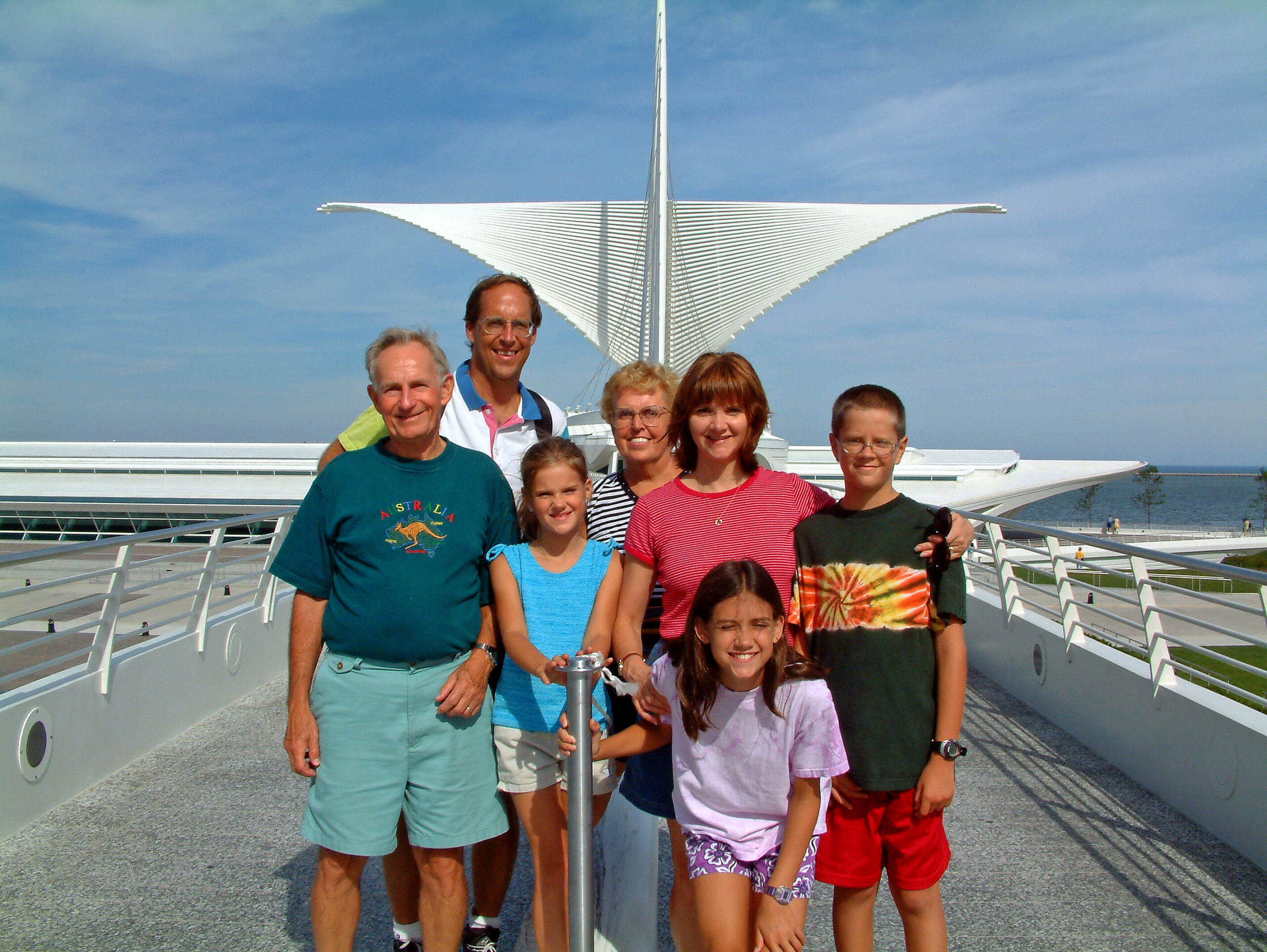Smiling family posing on a modern pedestrian bridge with a large white sail-shaped sculpture in the blue sky behind them.