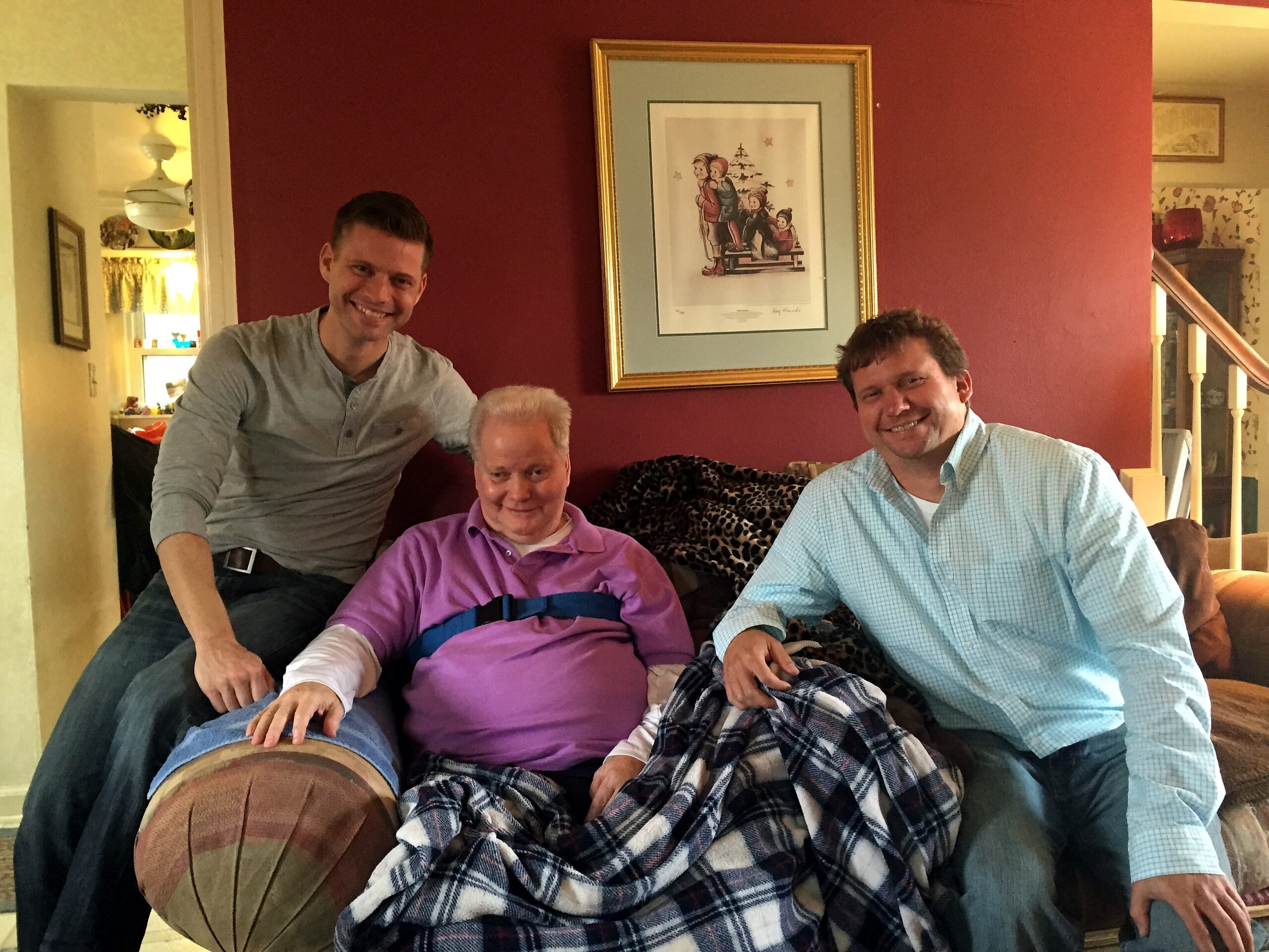 Three men sit close on a sofa in a cozy living room, smiling at the camera with a framed picture on the red wall behind them.