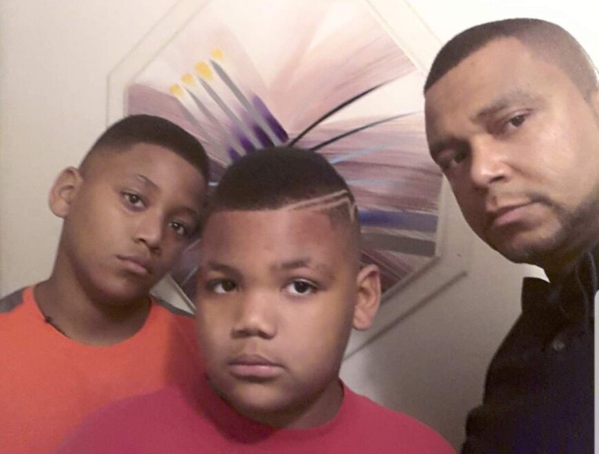 Three people pose for a close-up selfie indoors: a man on the right and two boys in red shirts, with the center boy showing a geometric hair design.