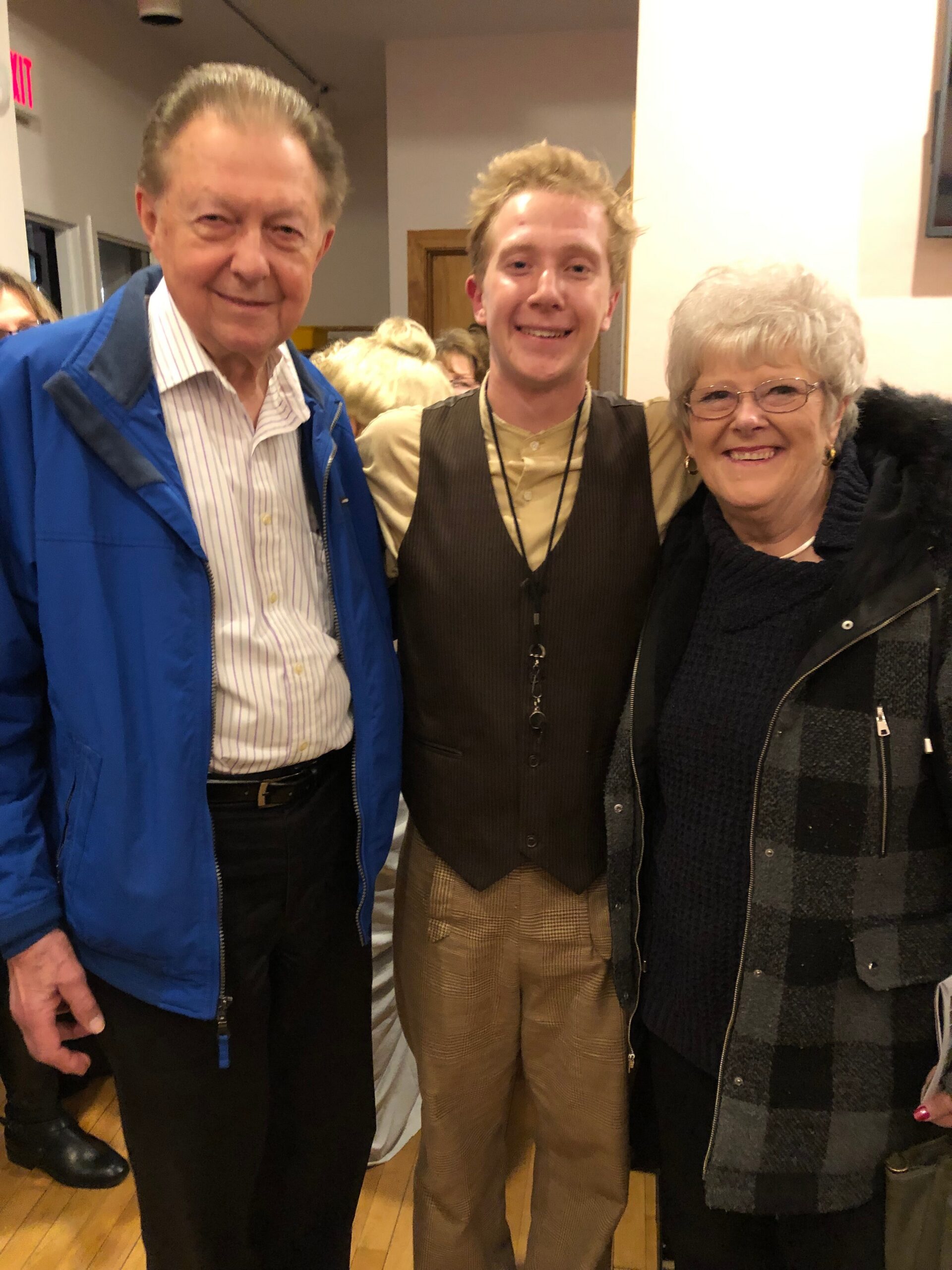 Three smiling people posing together indoors: an older man in a blue jacket, a younger man in a vest with a lanyard, and an older woman with glasses in a plaid coat.