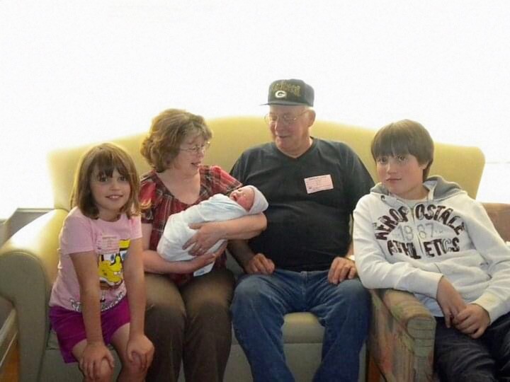 Family of five sits on a yellow couch: mother cradles a newborn wrapped in a blue blanket, with father and two older children nearby smiling.