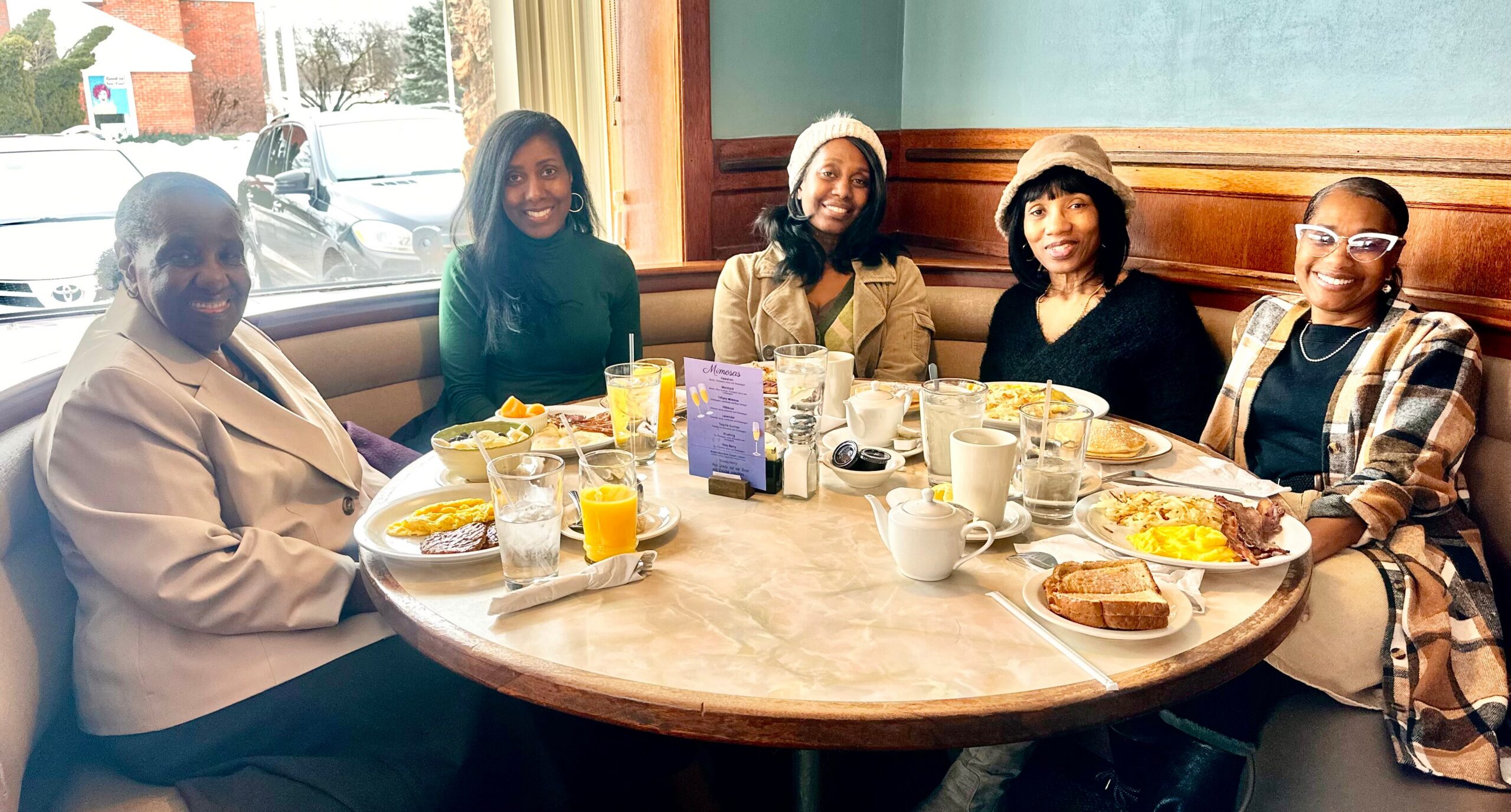 Five women smile and chat around a round table in a diner booth, with plates of breakfast and drinks visible.