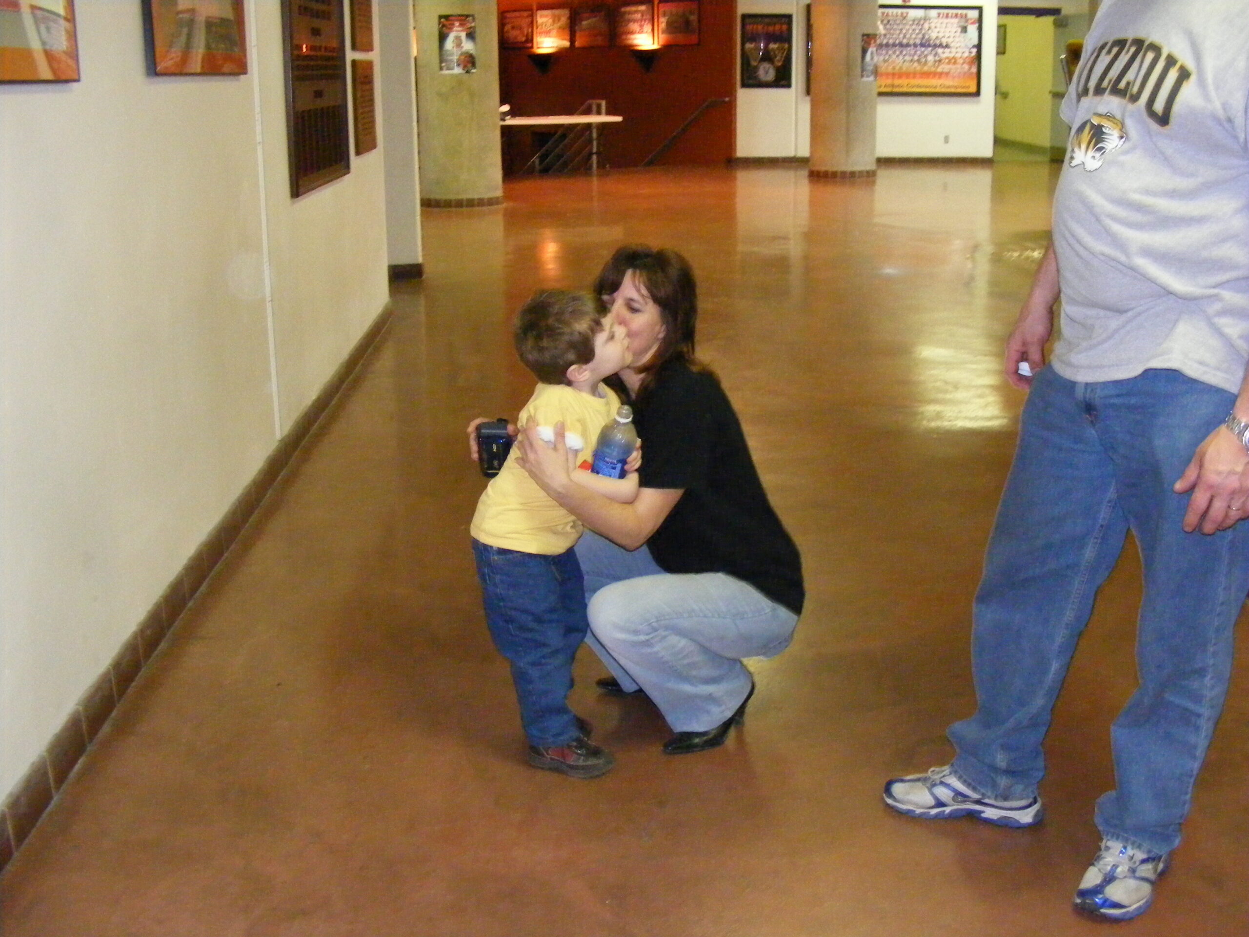 A woman kneels to hug and kiss a young boy who is holding a water bottle in a hallway.