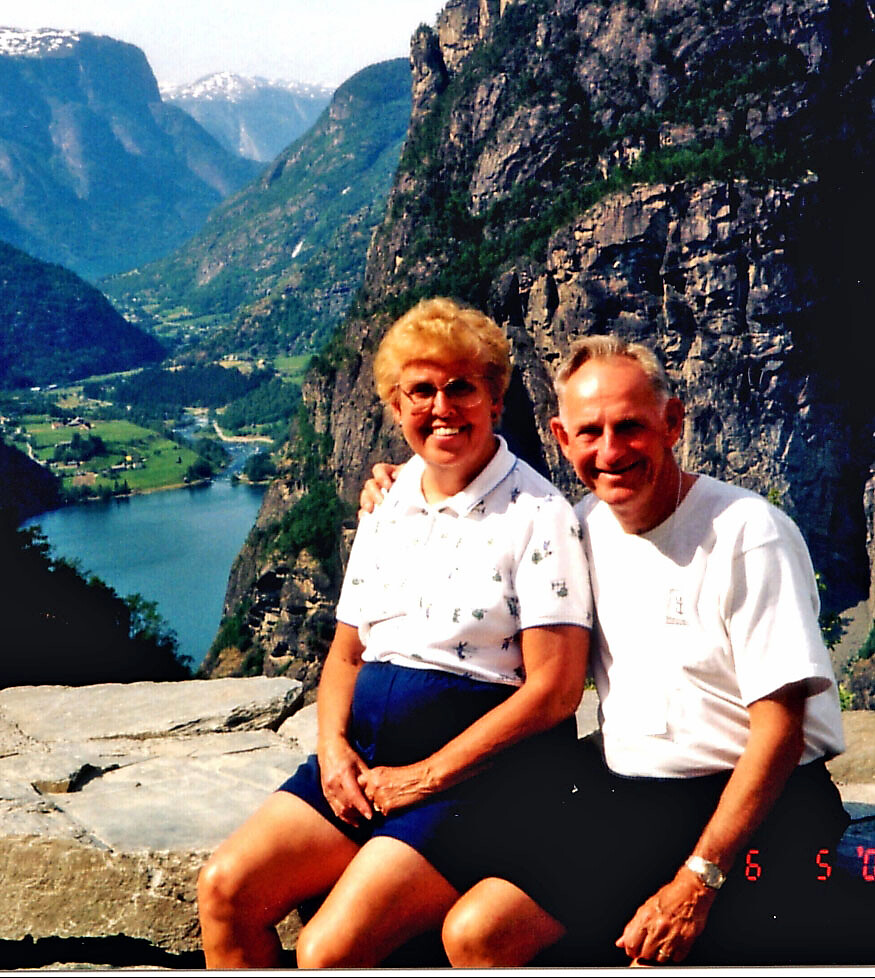 A smiling older couple sits on a rocky overlook with a deep valley, lake, and mountains in the background.