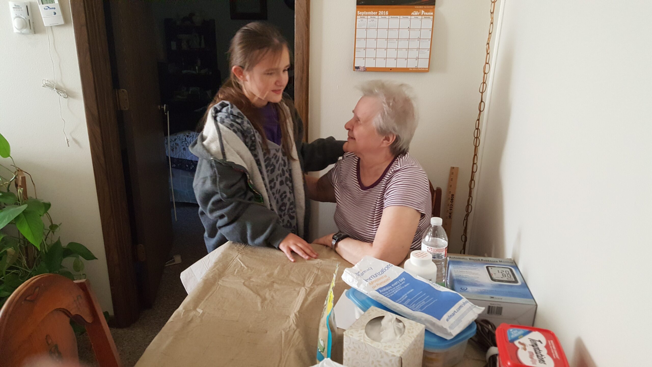 Young girl hugs an elderly woman seated at a table in a home, with a calendar on the wall nearby.