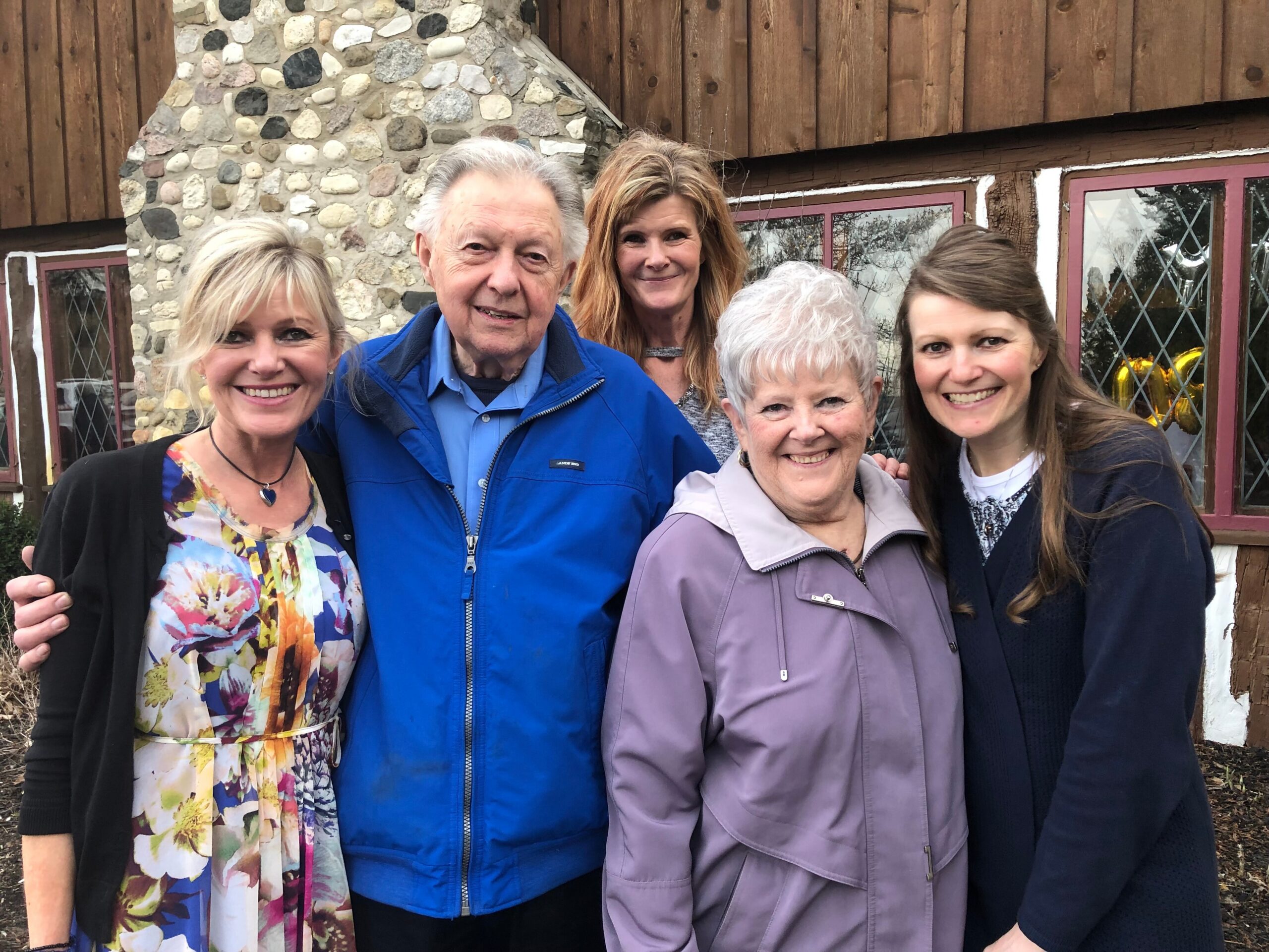 Group of five adults smiling at the camera outdoors in front of a stone chimney and wooden house.