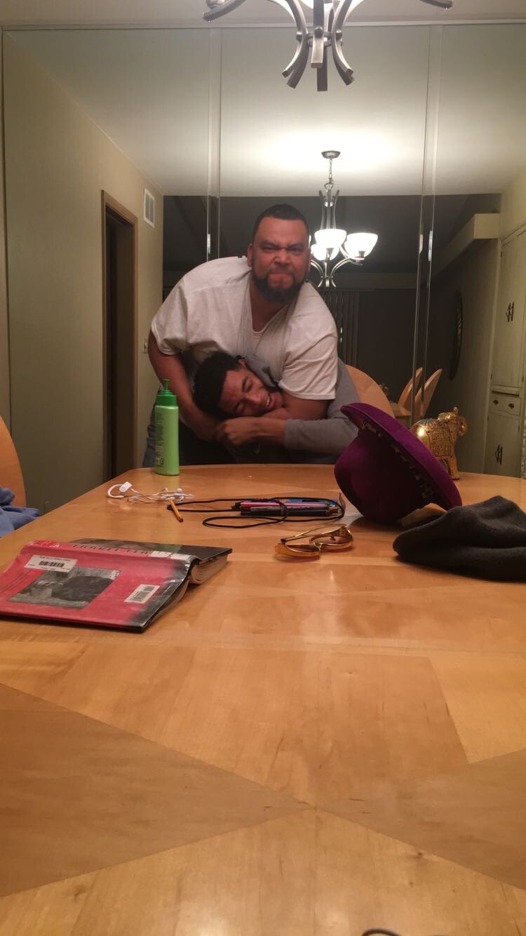 Two men wrestle or restrain each other at a dining table, one gripping the other with an intense expression; a green bottle and various items sit on the table in front of them.