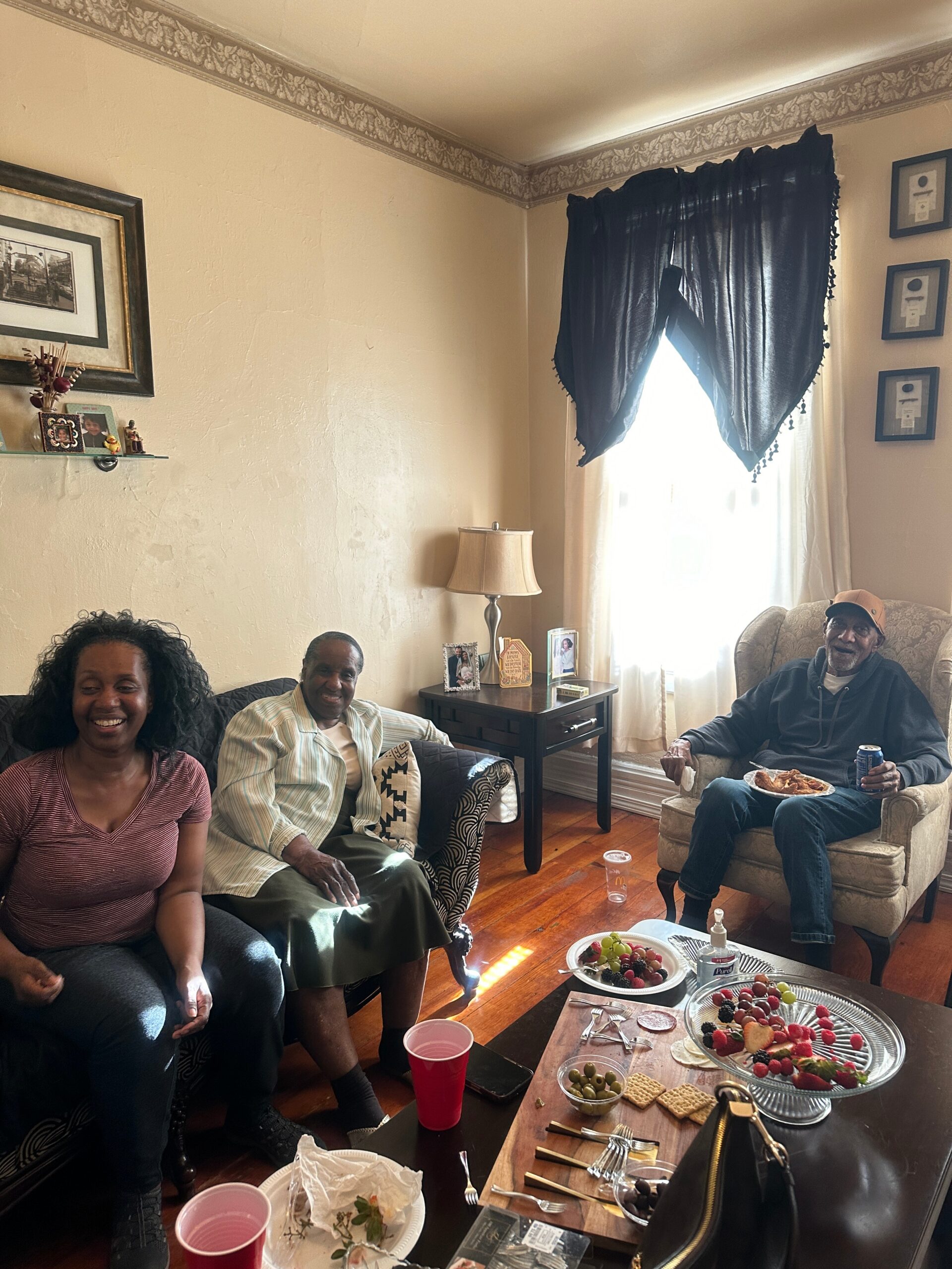 Three adults sit on couches in a cozy living room, smiling, with a snack-filled coffee table in front of them.