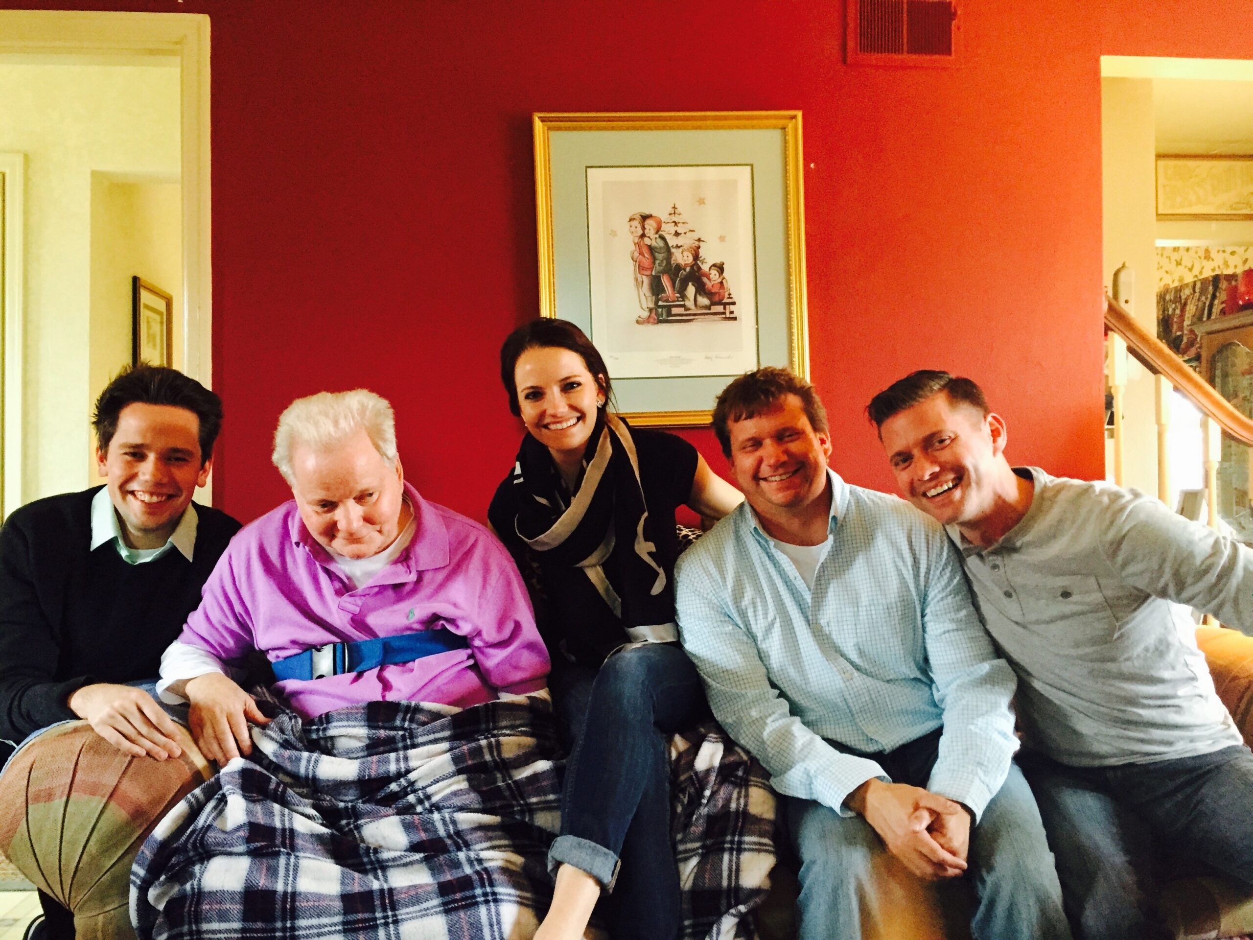 Five adults sit close together on a plaid blanket-covered sofa, smiling for a casual indoor group photo.