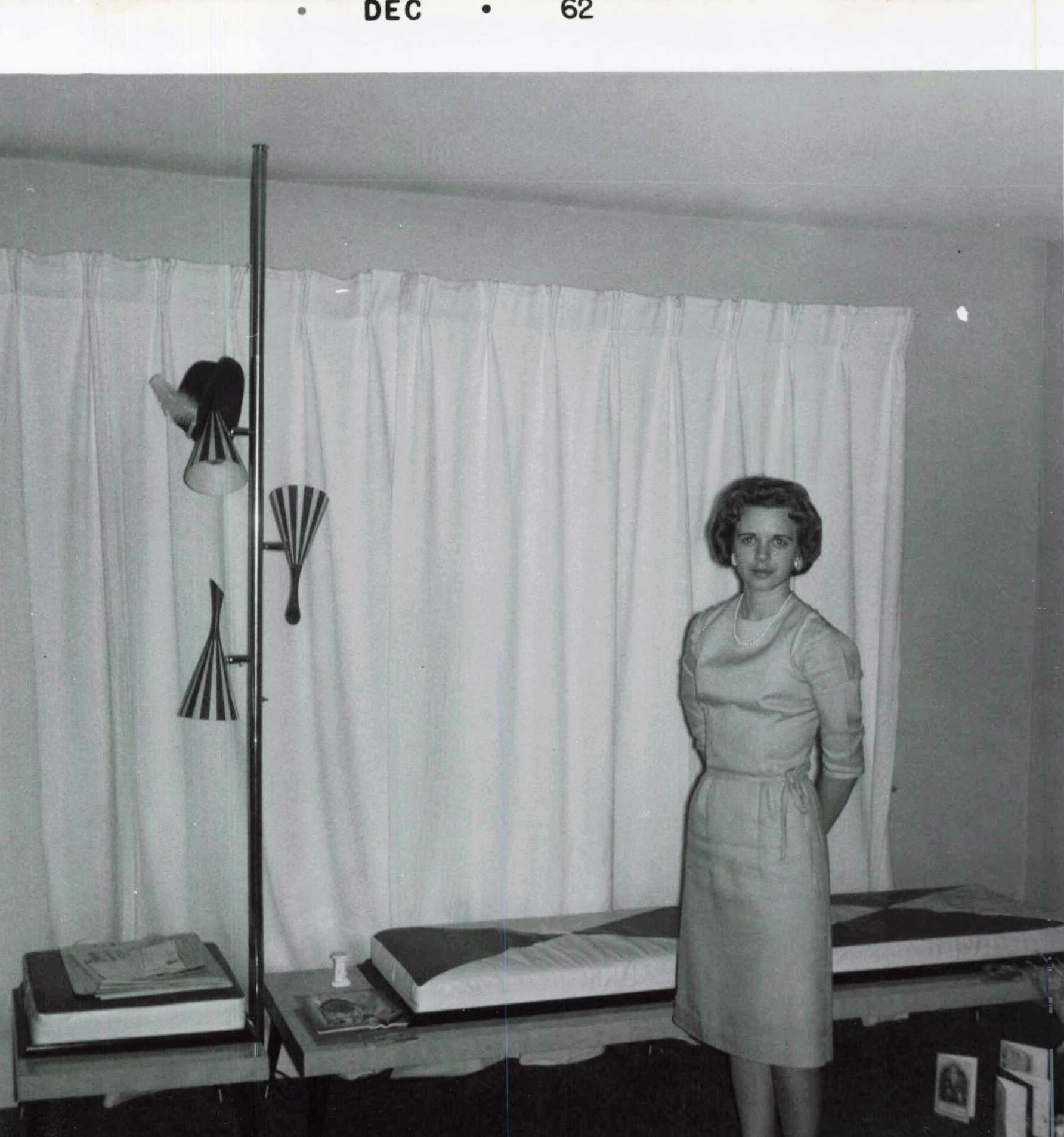 Woman in a vintage 1950s dress standing beside bed displays in a showroom, with a lamp and curtains in the background.