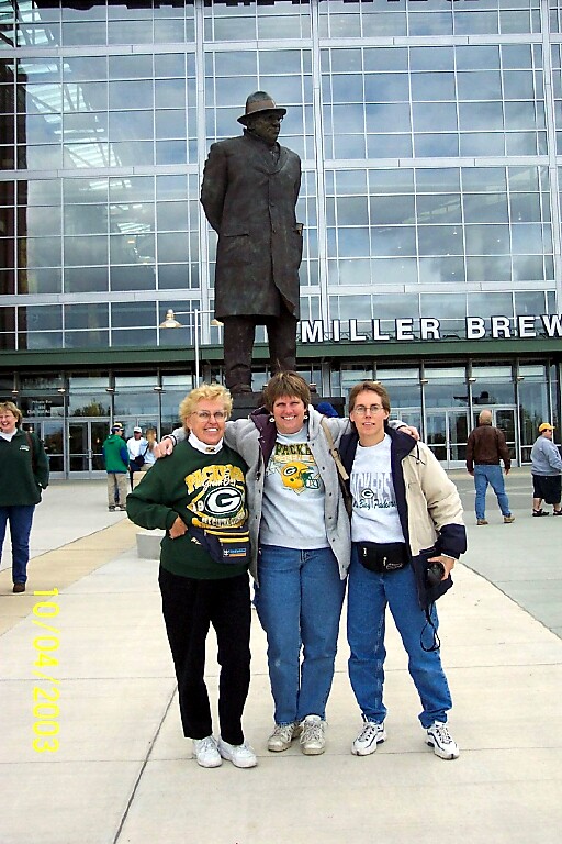 Three women pose with arms around each other in front of a tall statue outside the Miller Brewing Co. entrance; all wear Green Bay Packers shirts.