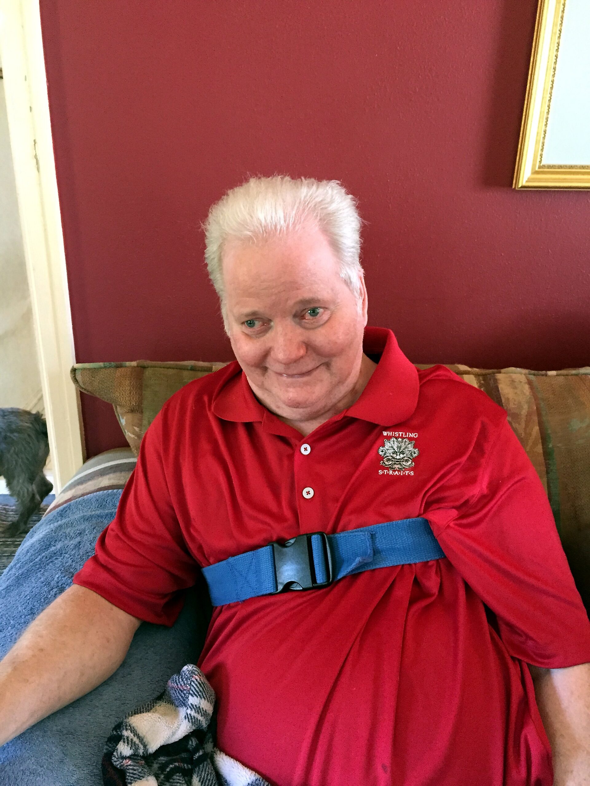 Older man with short white hair wearing a red polo shirt and a blue chest strap sits on a patterned sofa in a room with a maroon wall behind him.