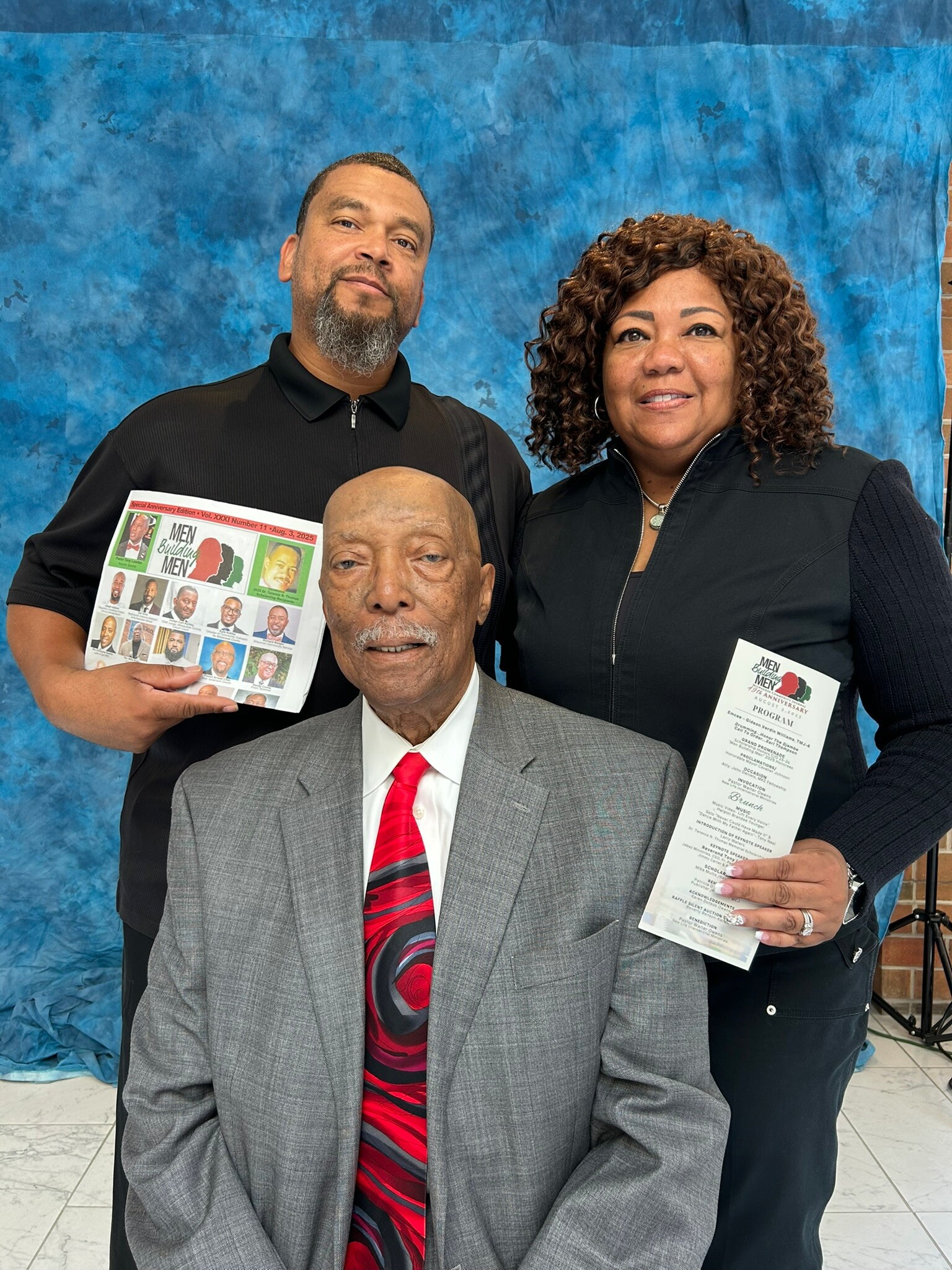 Three adults posing for a photo against a blue backdrop: a man in a gray suit seated front, flanked by a man holding a magazine and a woman holding a program, all smiling.