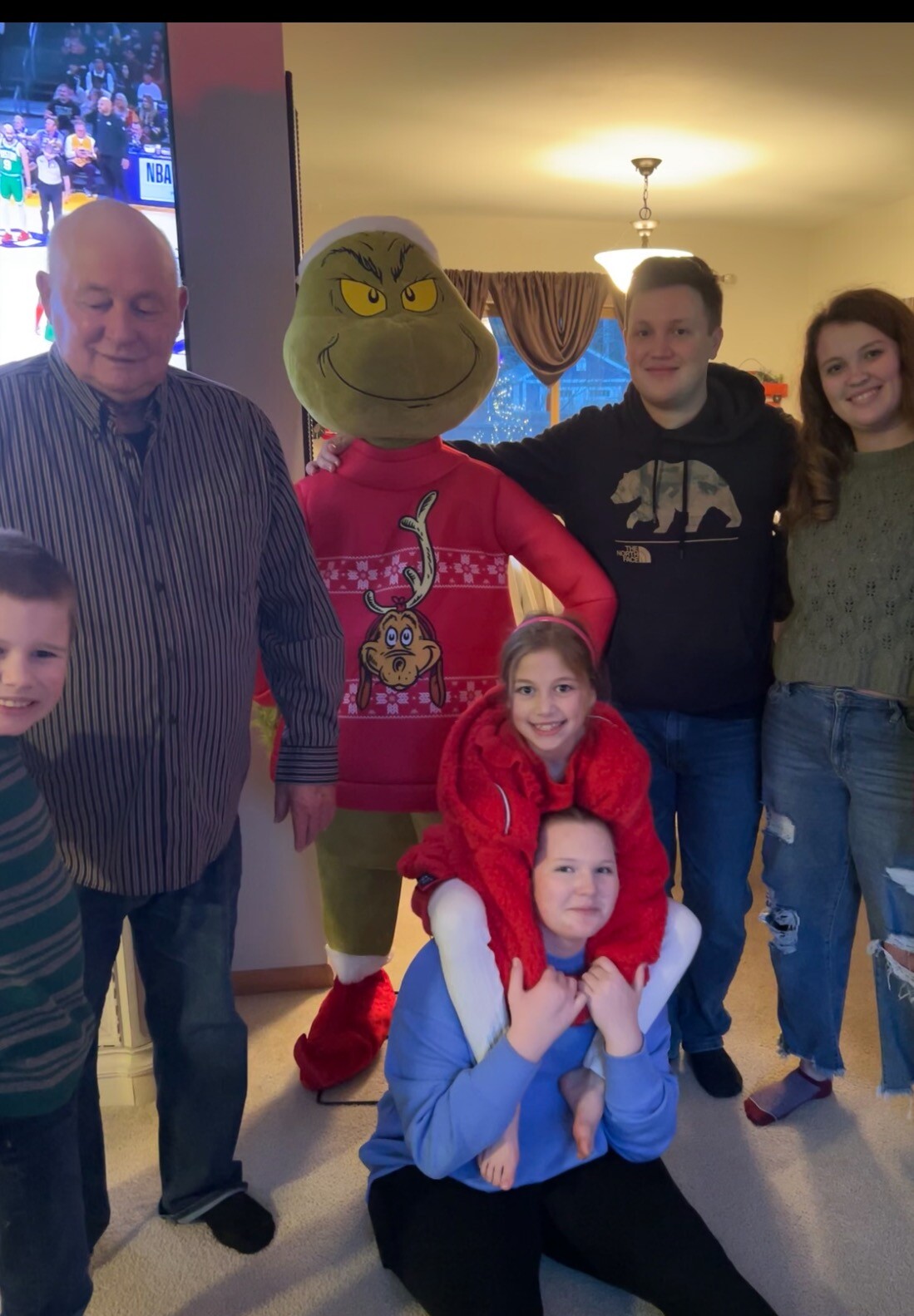 A group of people posing with a Grinch mascot indoors, all smiling for the photo in a cozy living room setting.