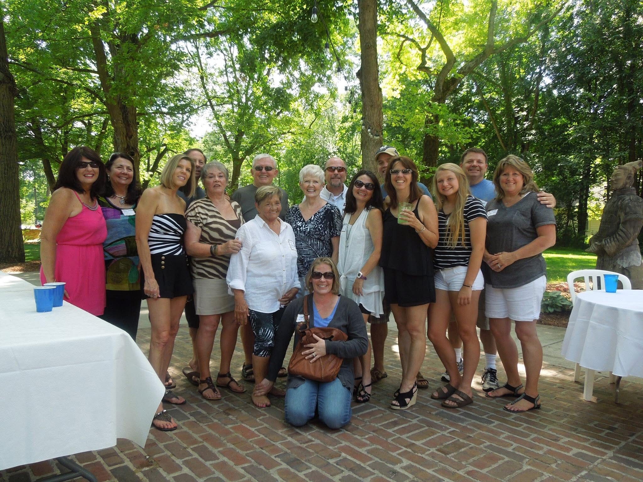 Group of adults posing together outdoors under trees at a sunny gathering, smiling at the camera in a park setting with tables nearby.