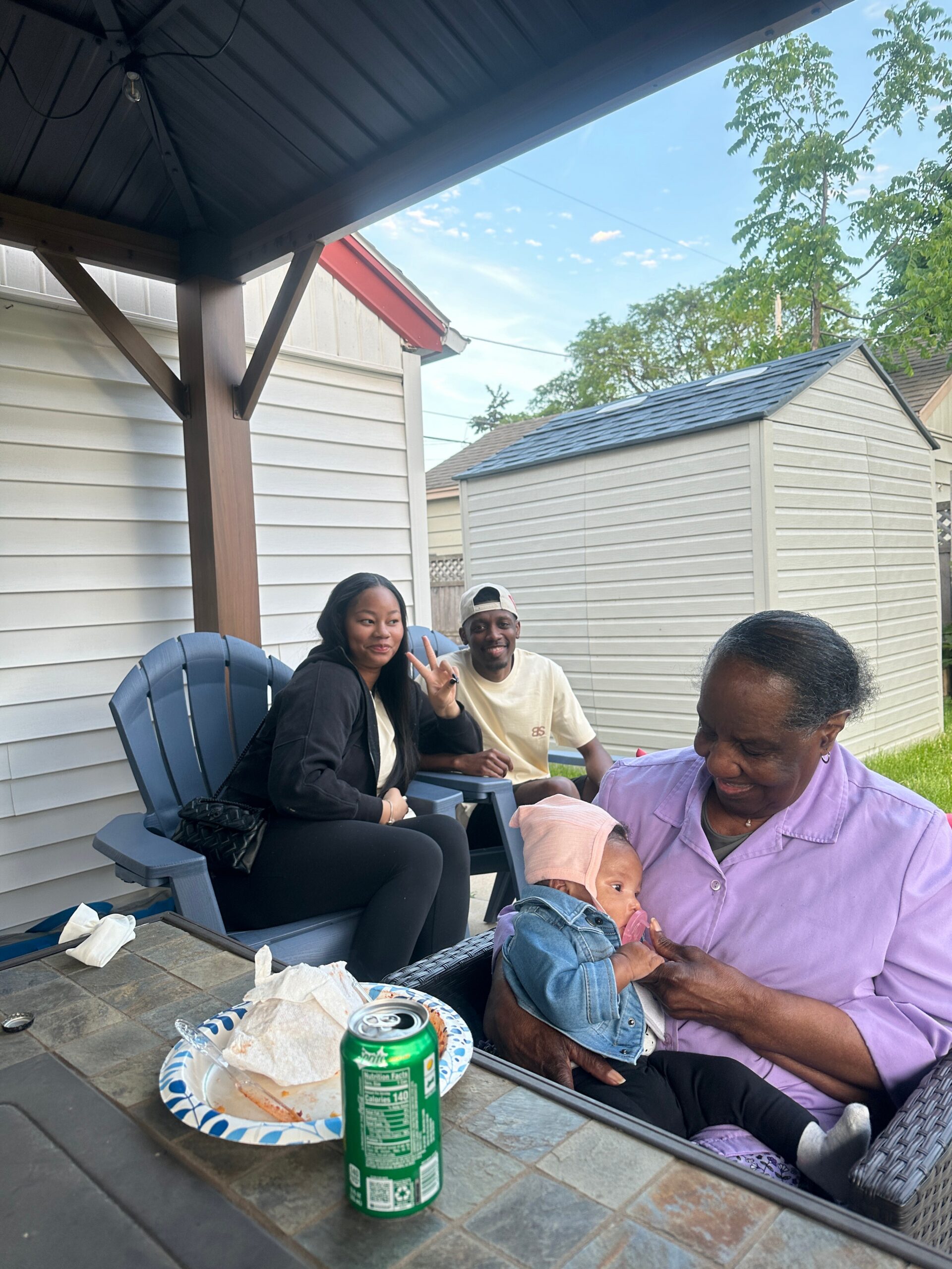 A group of people sitting on a porch with a baby, snacks and drinks on a tiled table nearby, outdoors with sheds in the background (aluminum siding).