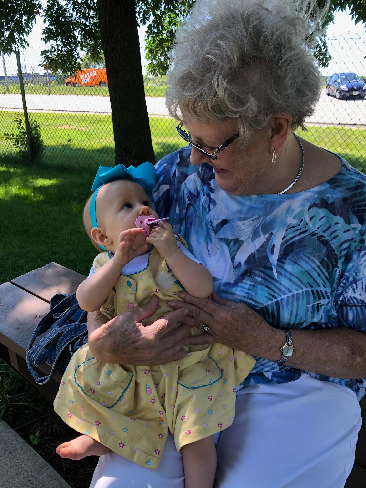 Older woman with short gray hair in a blue patterned top holding a baby girl on a park bench outdoors, the baby with a pink pacifier and turquoise bow headband.