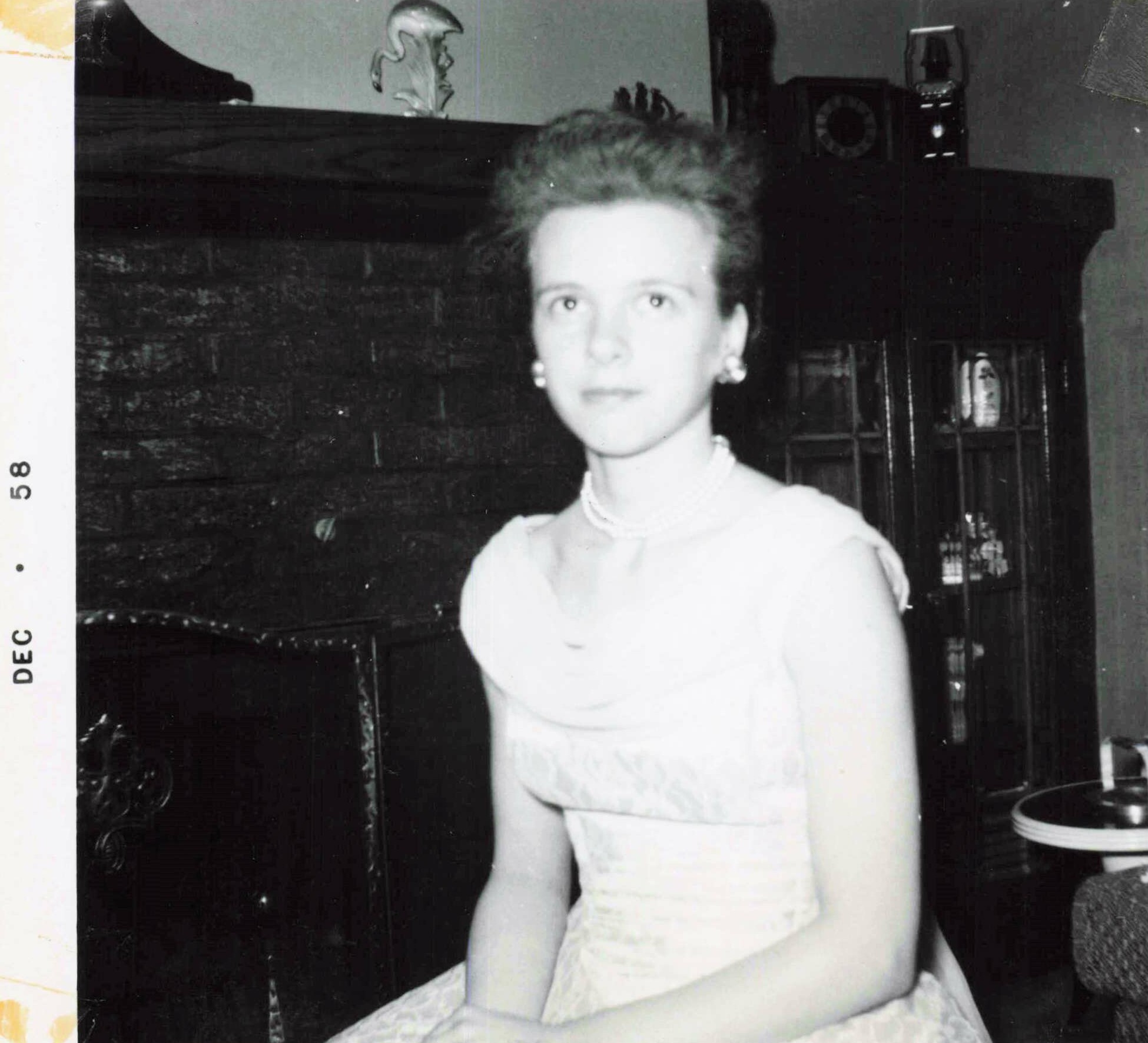 Black-and-white photo of a woman in a light dress with pearls, seated in a living room in front of a dark fireplace and cabinet.