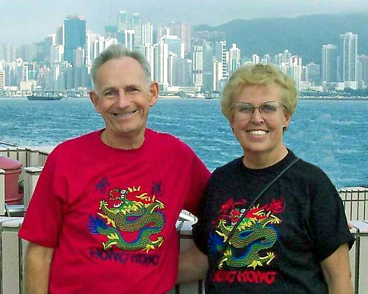 Smiling older couple posing by a marina with a city skyline and distant mountains in the background.