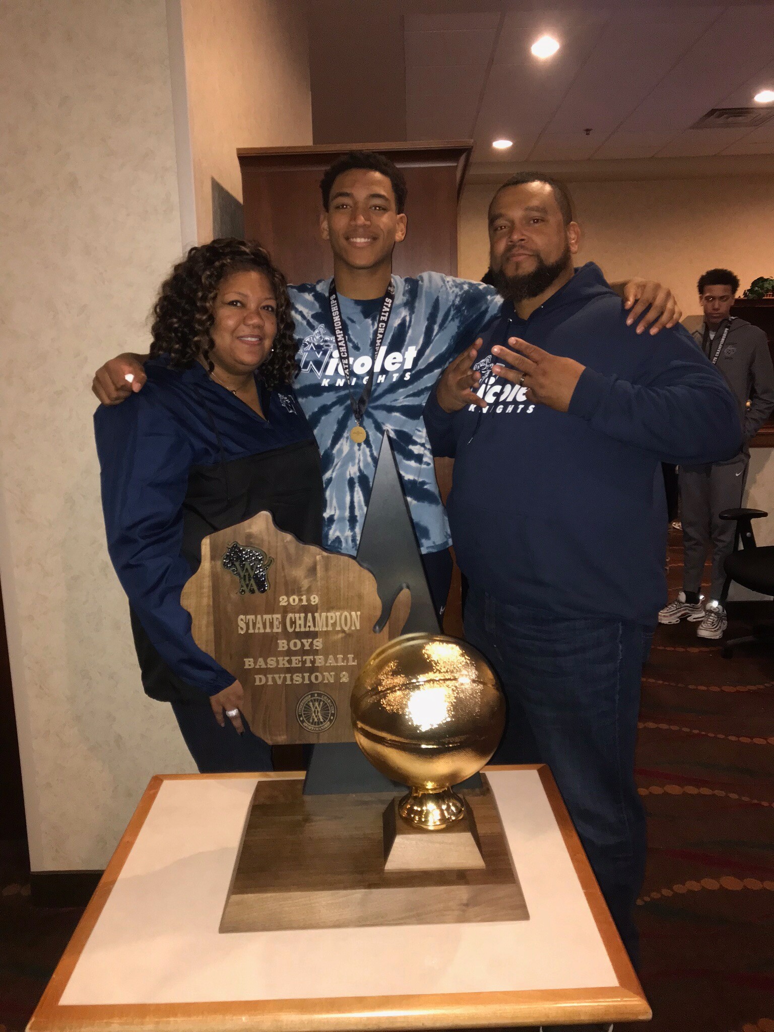 Three people pose together with a 2019 State Champion Boys Basketball Division 2 trophy plaque, celebrating a win.