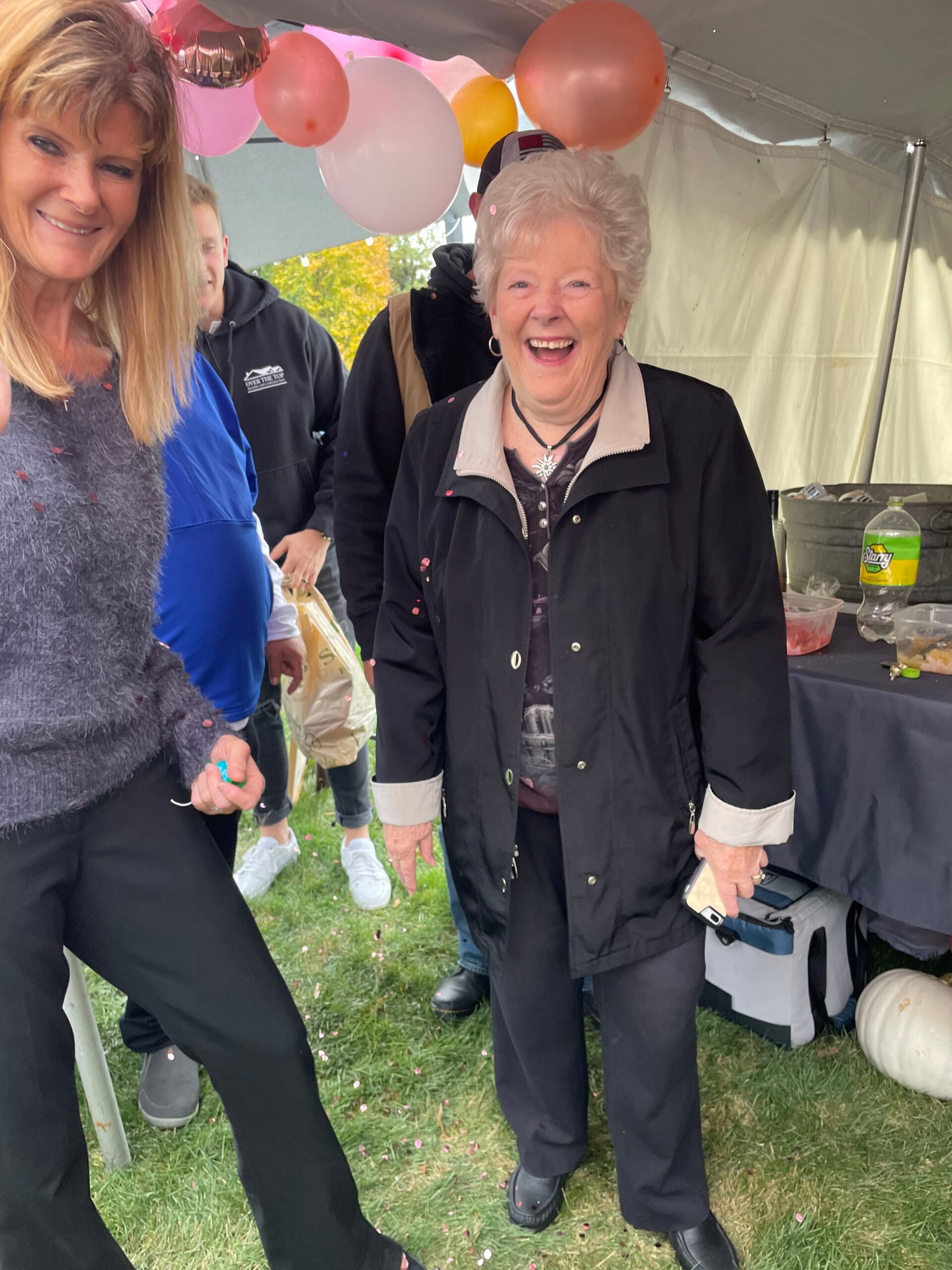 Cheerful older woman in a black coat at an outdoor celebration under a tent, with balloons in the background.