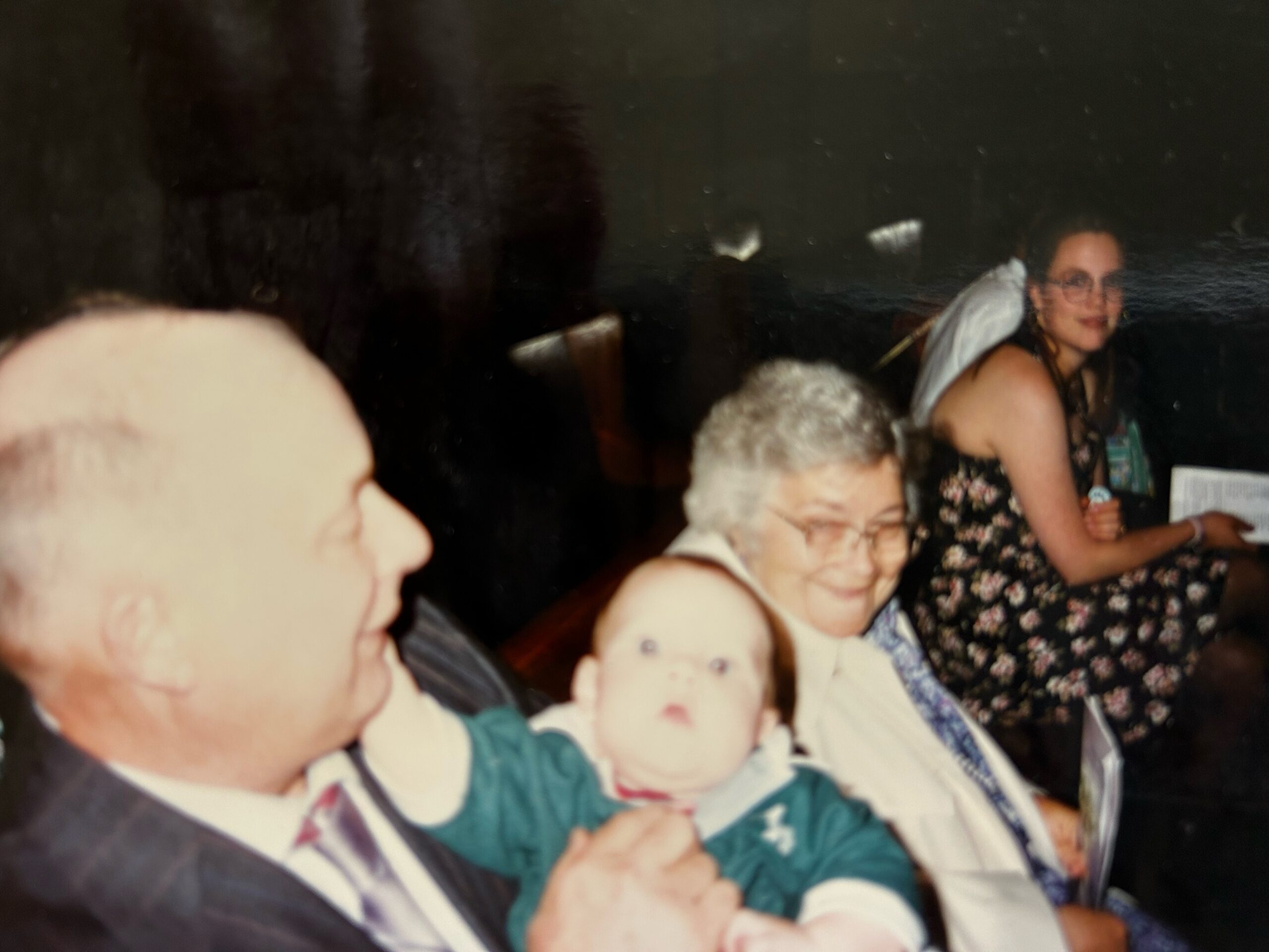 People at a crowded celebration, a baby being held in the foreground near adults smiling at the camera in a dimly lit room