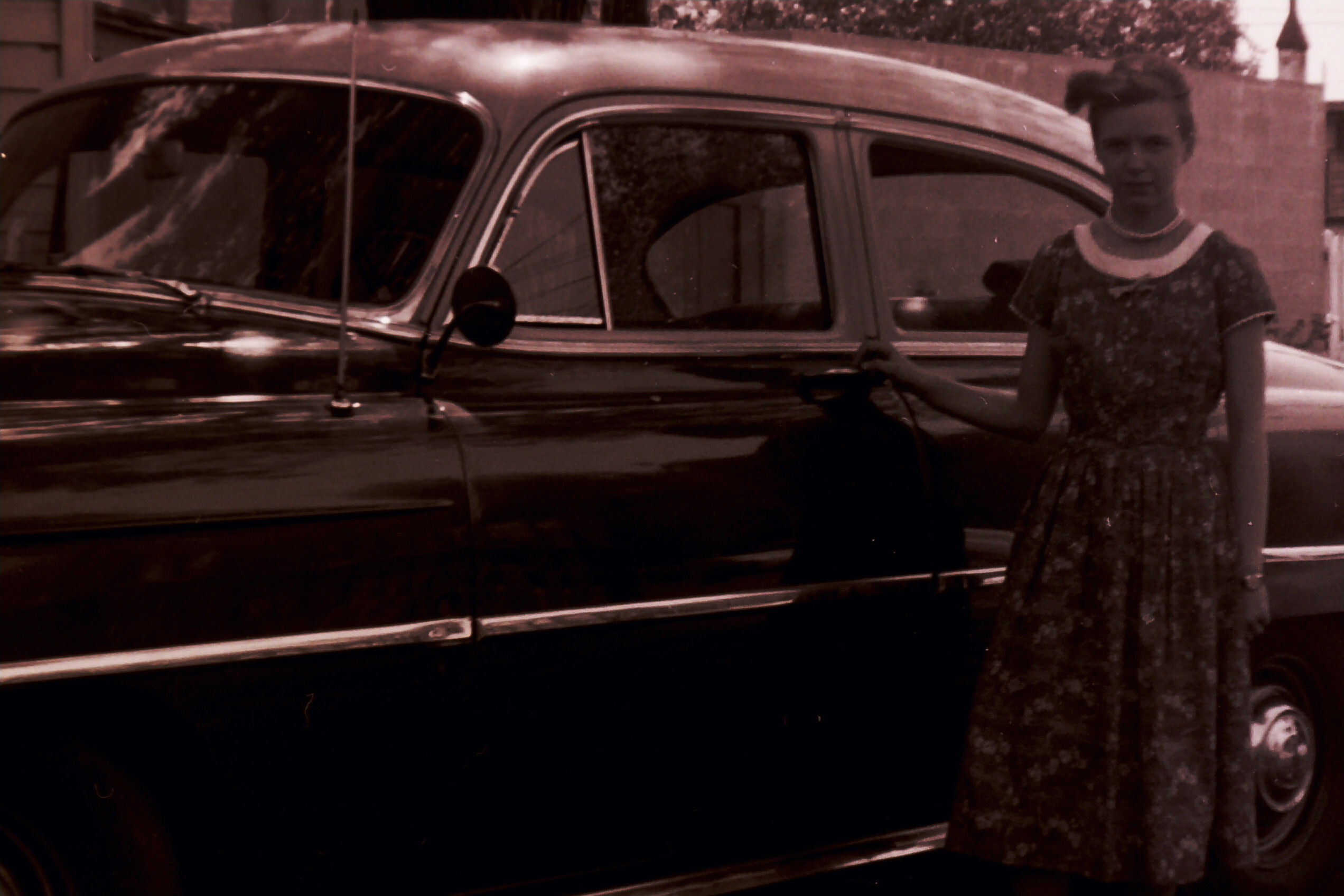 A woman in a vintage patterned dress stands beside a dark classic car outdoors.