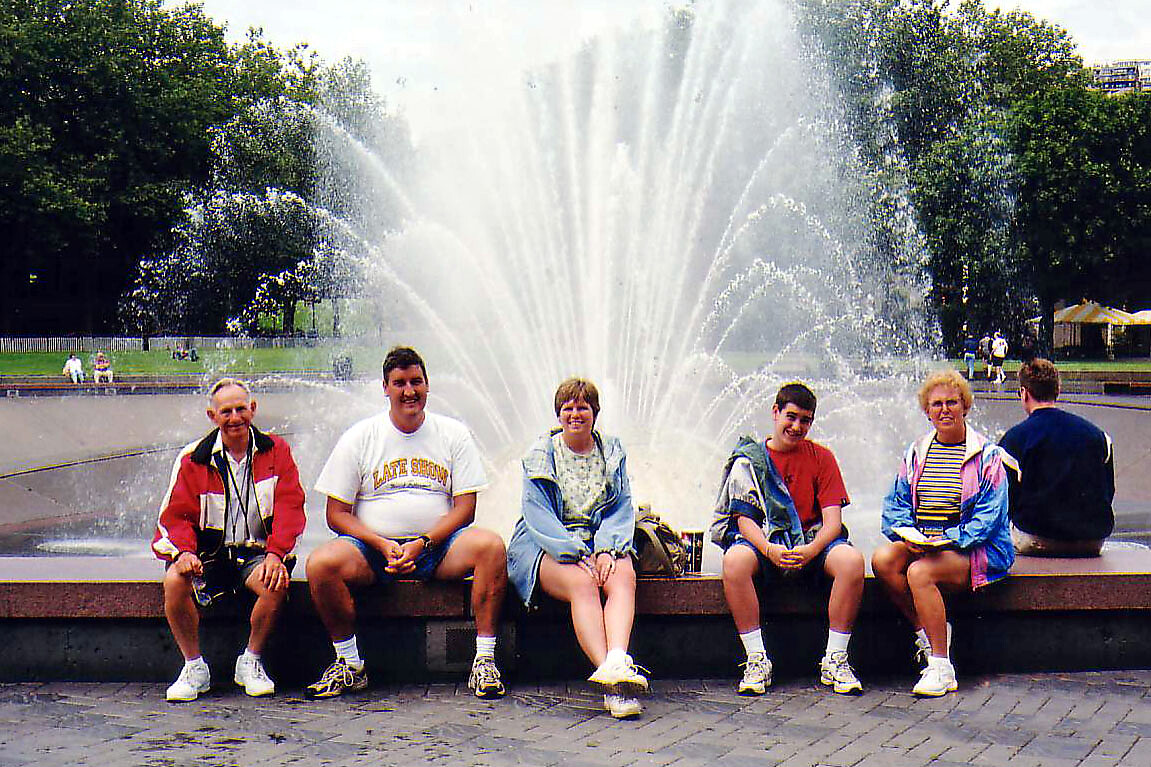 Five people sit on the edge of a large park fountain, smiling at the camera with water jets spouting behind them.