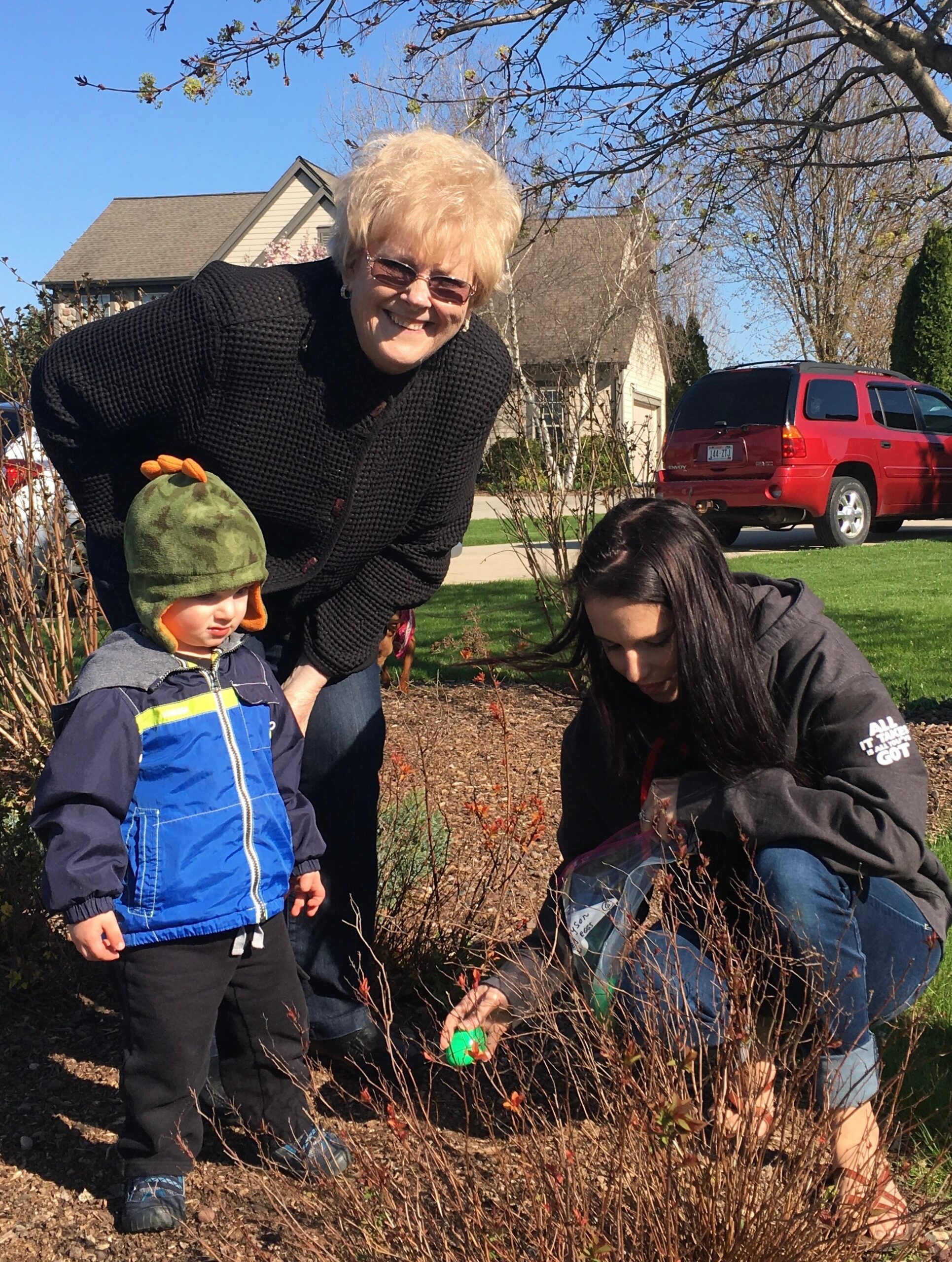Smiling older woman bending beside two children while planting in a front yard; bare shrubs and a red SUV in the background on a sunny day.