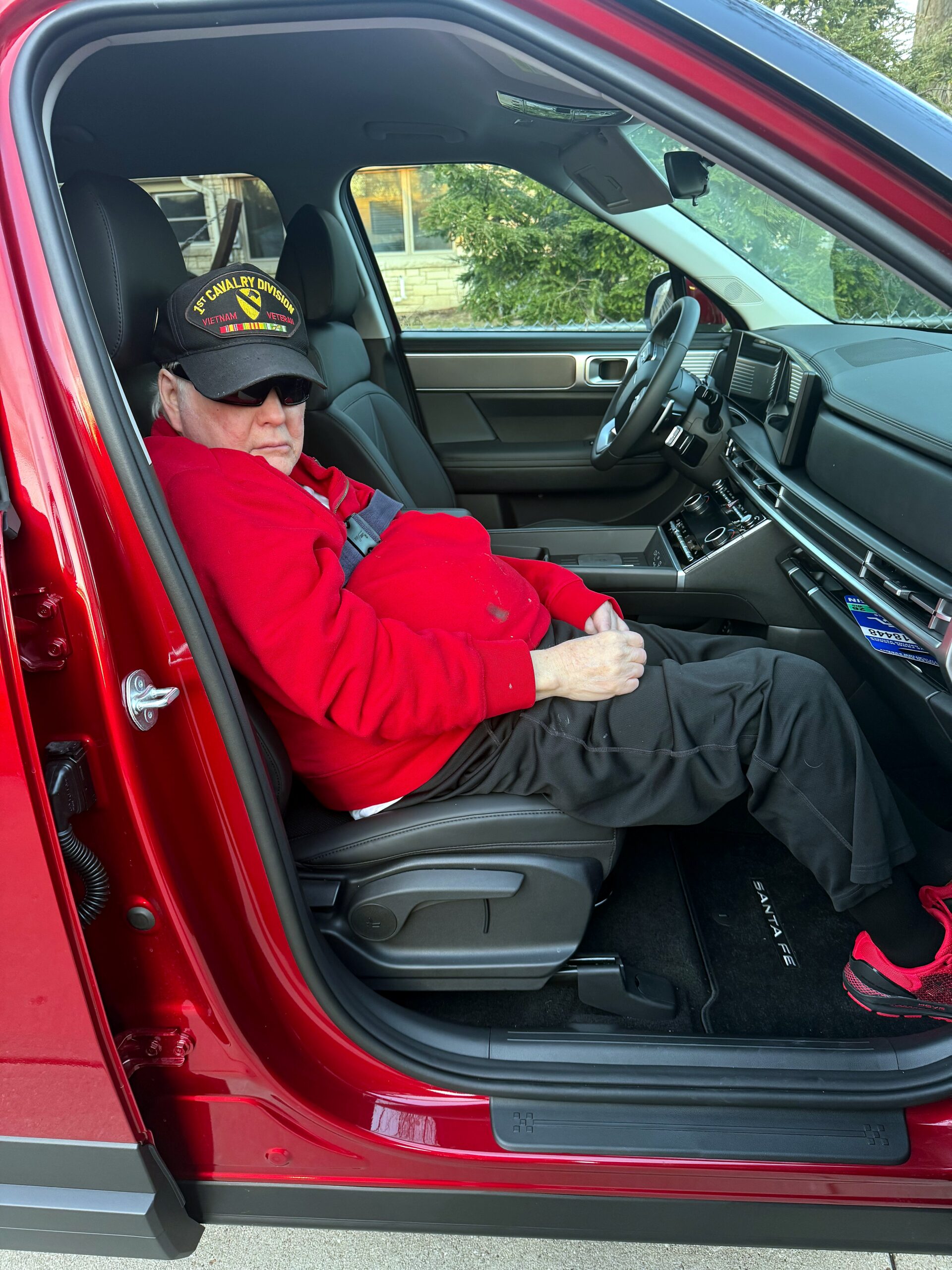 Older man wearing a red jacket and a black cap with '1st Cavalry Division Vietnam Veteran' sits in the driver's seat of a red vehicle, looking ahead.