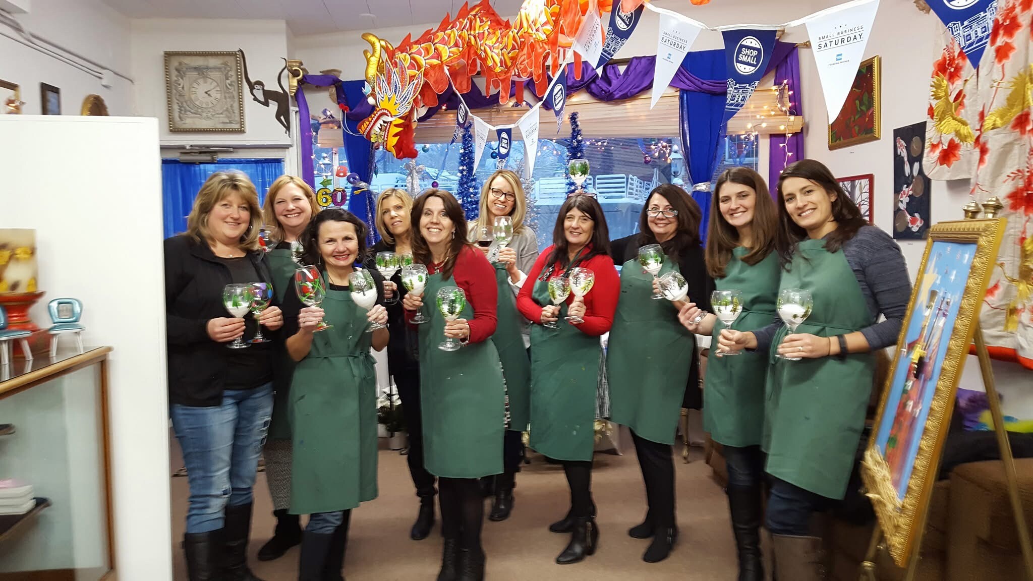Group of women in green aprons posing with wine glasses inside a festively decorated shop, smiling at the camera.