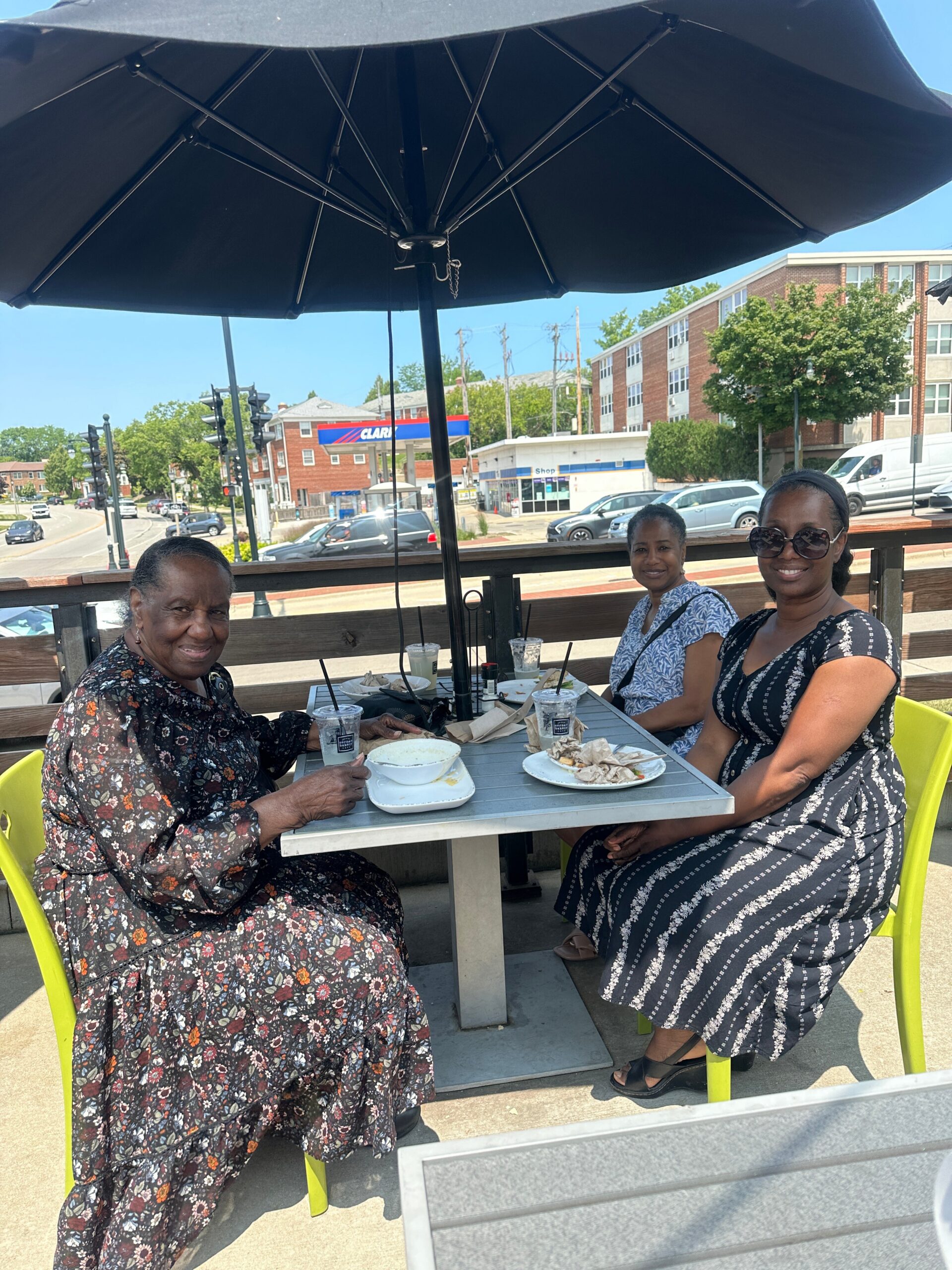 Three women sit at an outdoor cafe table under a large black umbrella, enjoying drinks and food on a sunny street.