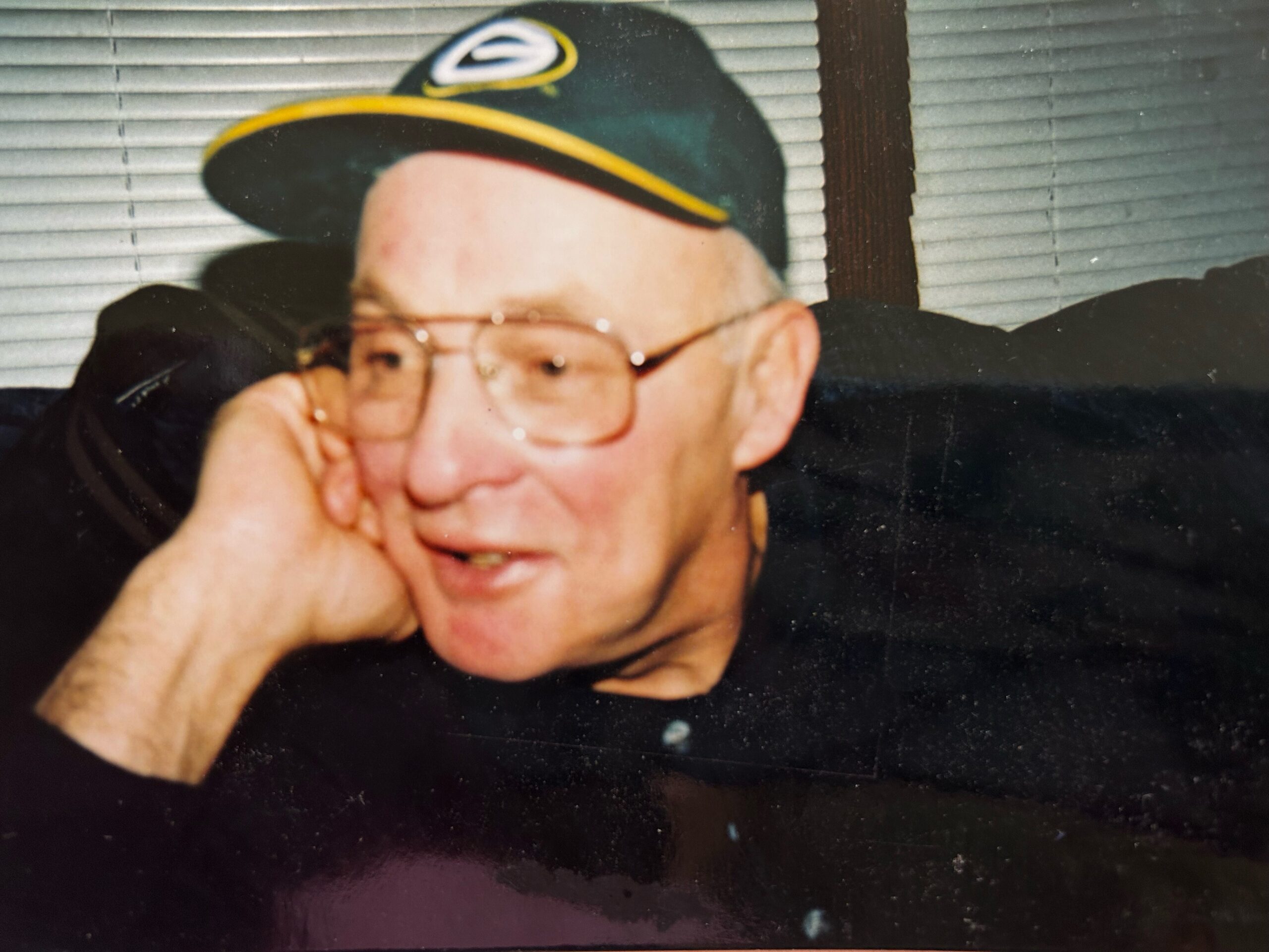 Close-up of an older man wearing glasses and a dark cap with a yellow brim, resting his chin on his hand in front of window blinds.