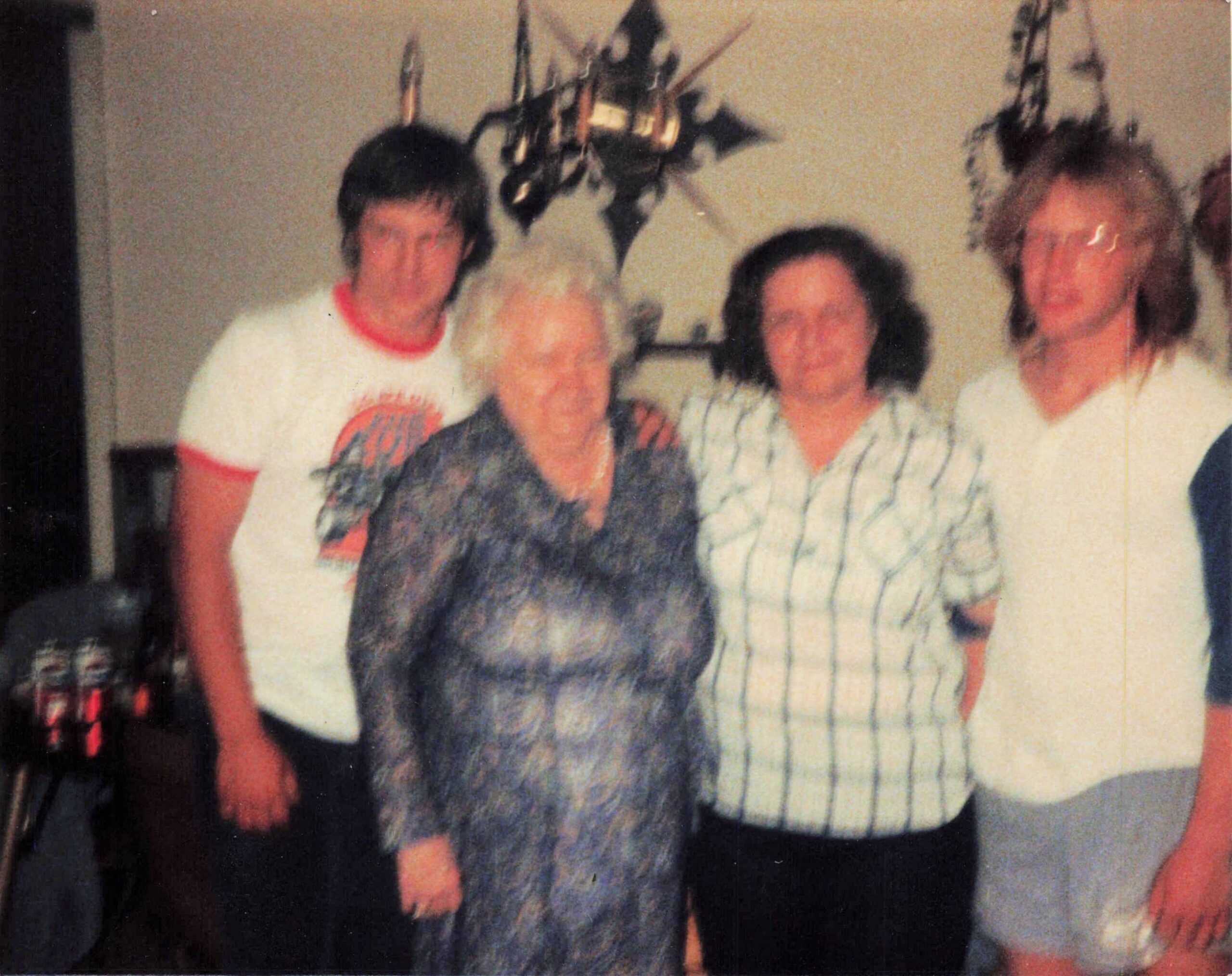 Three adults posing together indoors for a casual group photo; a woman in a patterned blouse sits center with two men beside her, background shows hanging decor.