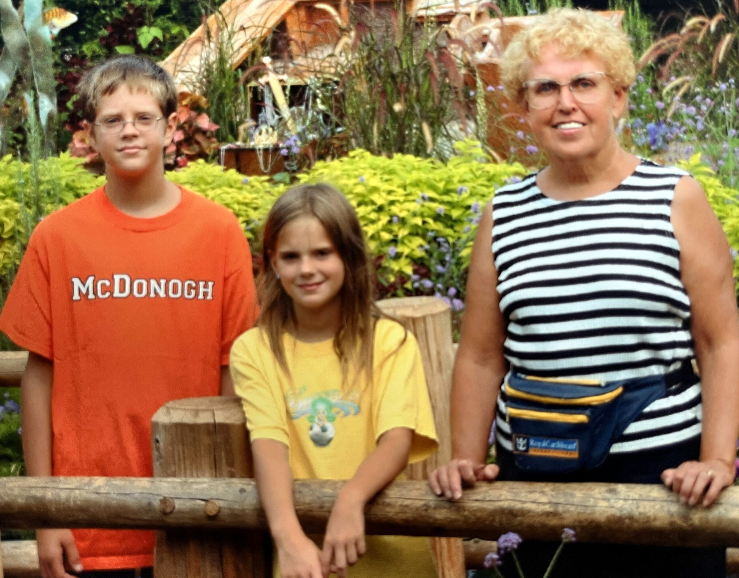 Three people pose behind a wooden railing in a garden with yellow-green shrubs and purple flowers.