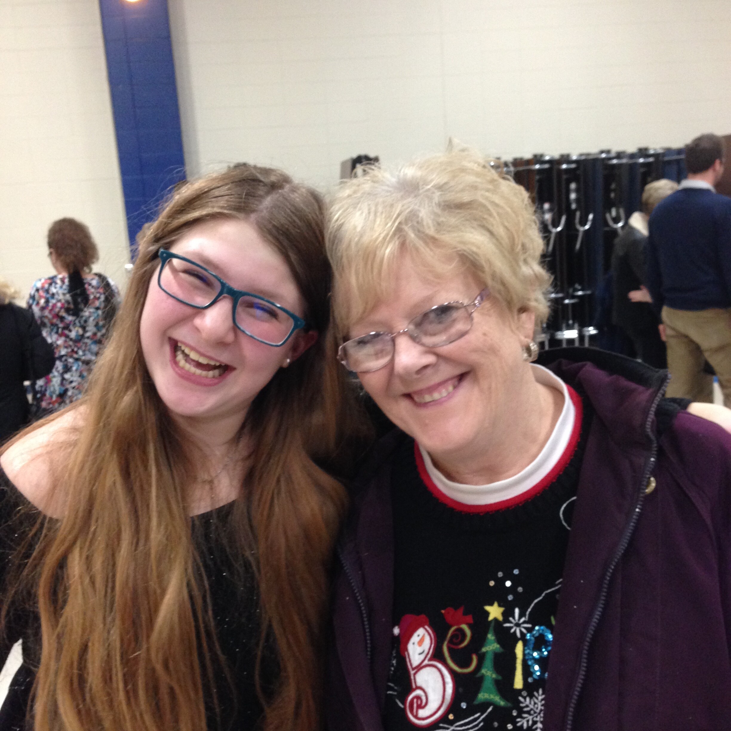 Two women smiling for a selfie in a school gym, wearing festive clothing and glasses, with coats on racks in the background.