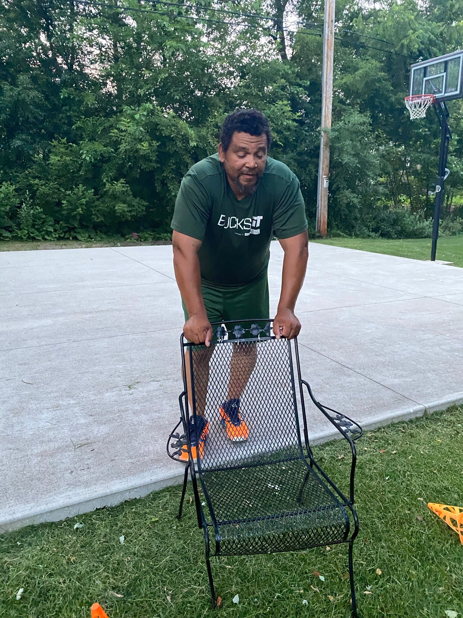 Man in a green shirt and shorts leaning on a metal outdoor chair on a concrete basketball court, with a hoop in the background.