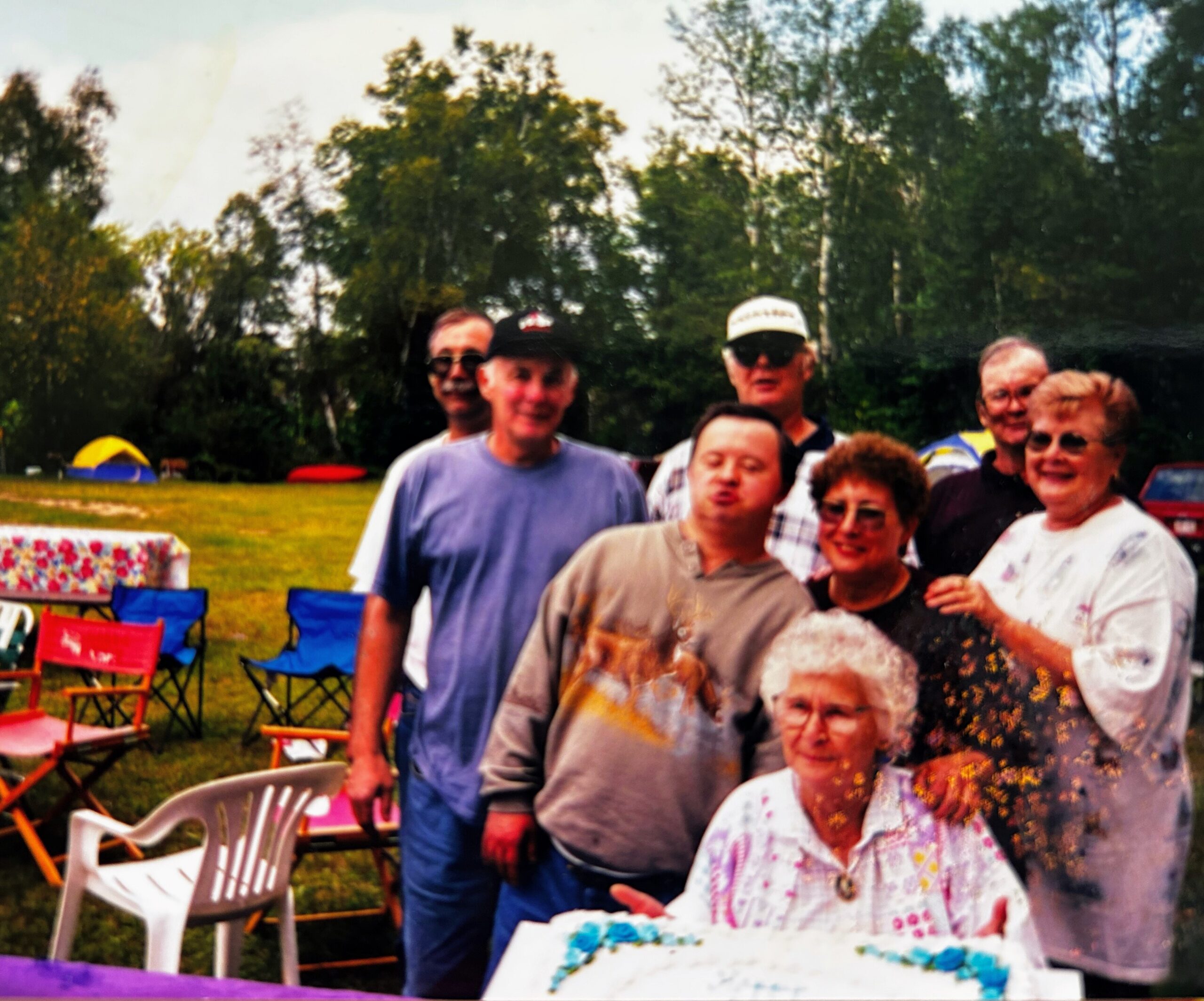 Group of smiling adults and children posing outdoors at a sunny gathering with colorful chairs and a painted artwork in the foreground.