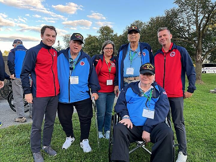 Group of veterans wearing red and blue jackets, smiling outdoors on grass with trees in background; some wear lanyards and caps, one in a wheelchair in front.