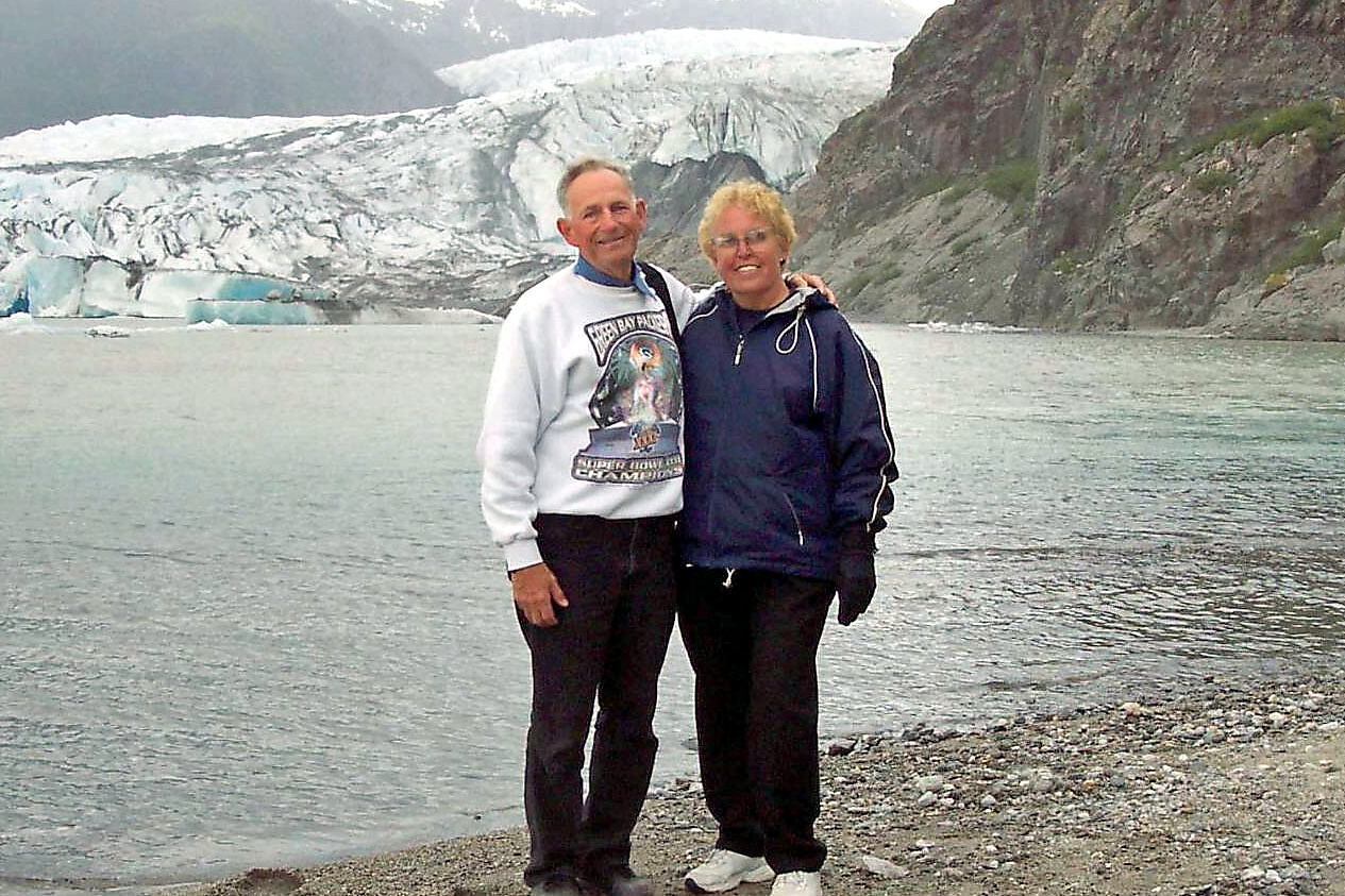 A couple stands arm-in-arm on a pebbled shore, with a glacier and rocky cliffs in the background.