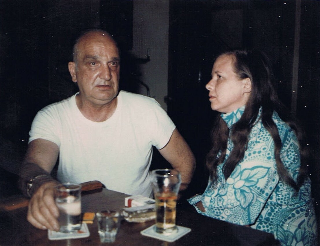 An older man in a white t-shirt and a woman in a teal patterned blouse sit at a dimly lit table with drinks and a pack of cigarettes between them.