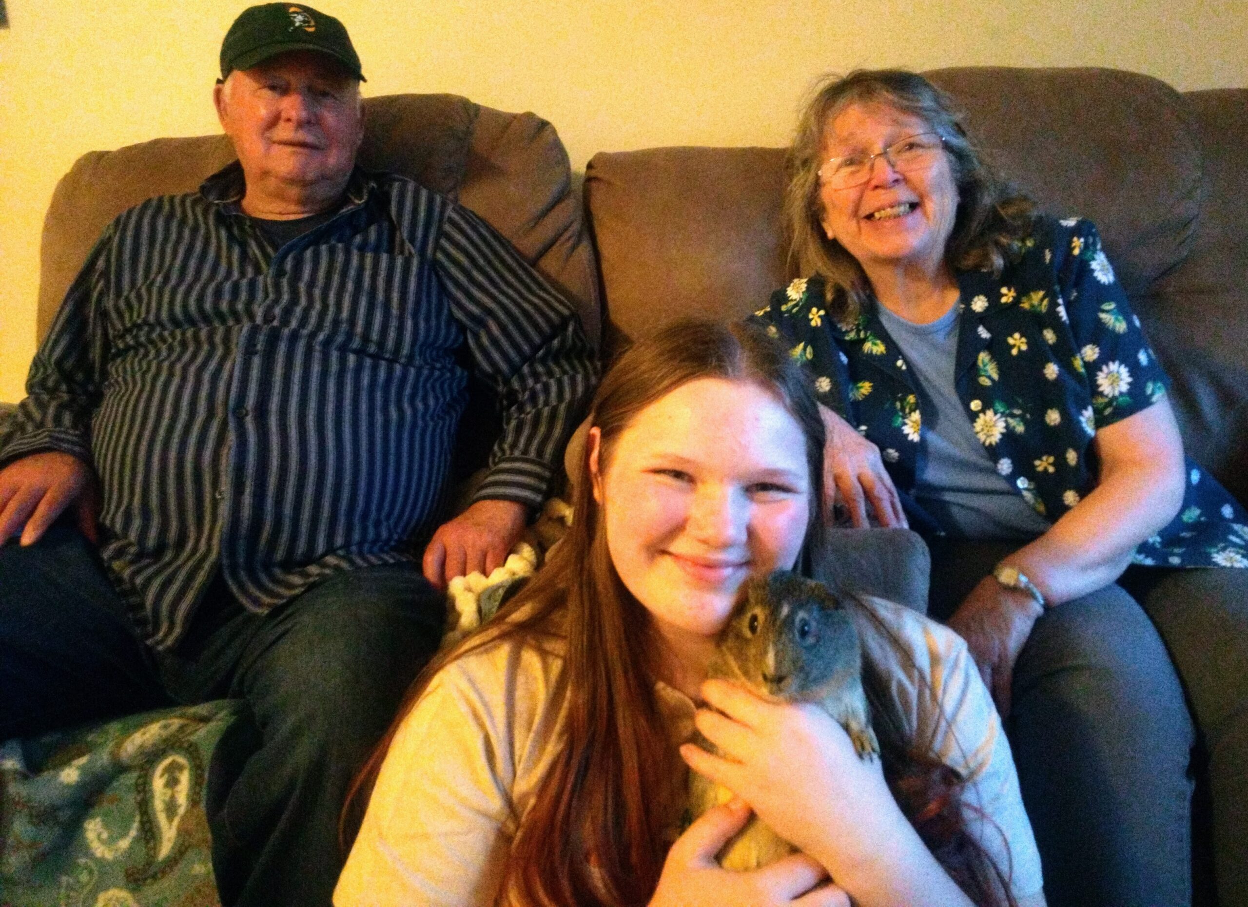 Three people sit on a brown couch: a man in a striped shirt and cap on the left, a smiling woman in a floral shirt on the right, and a girl in front holding a small gray pet close to her chest.