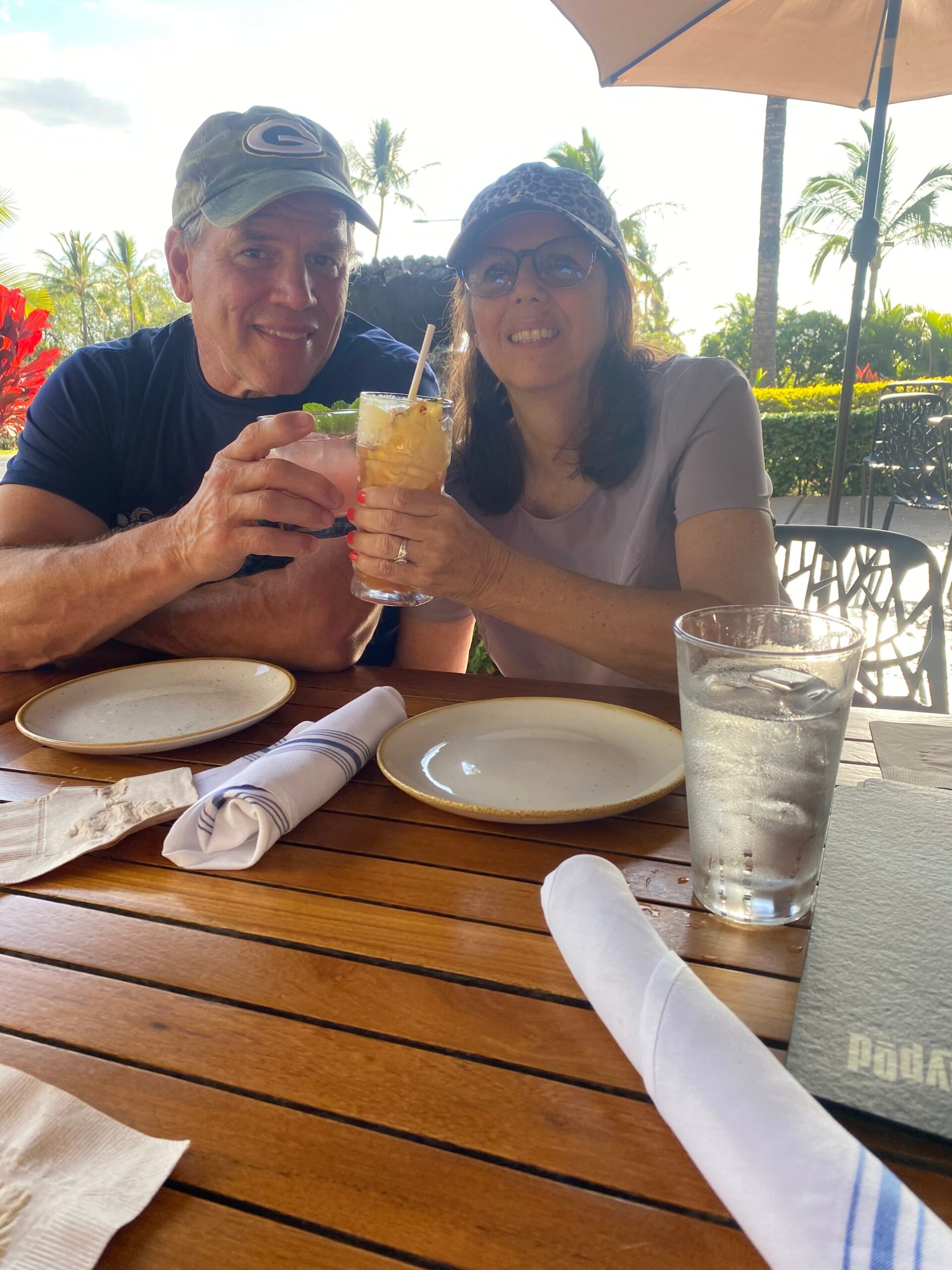 Two people at an outdoor seaside restaurant toast with colorful drinks, smiling at the camera, palm trees in the background.