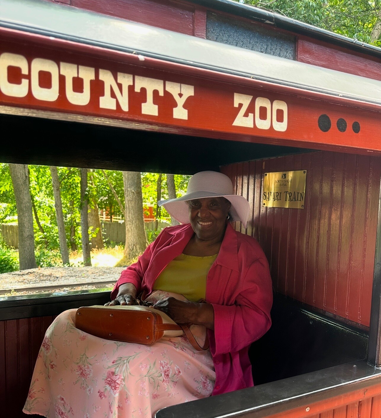 Smiling woman in a pink jacket and white hat sits inside a red safari train car at the zoo with a floral dress and purse on her lap.