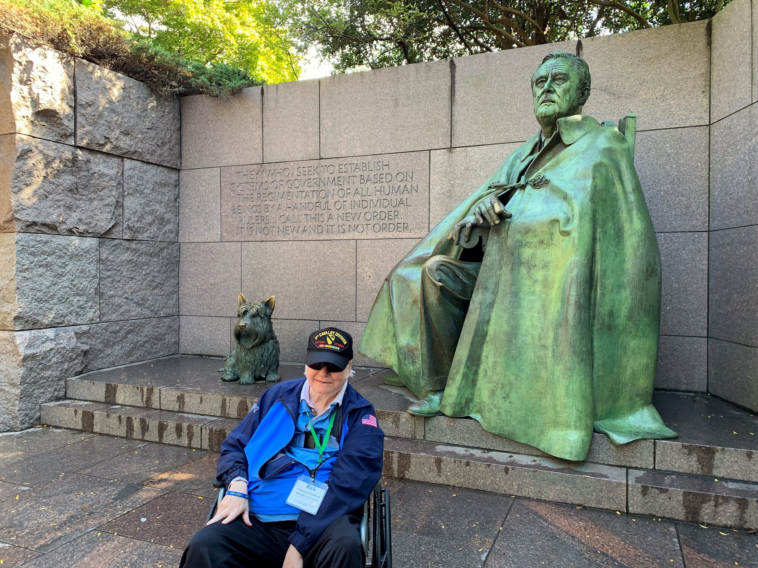Person in a wheelchair wearing a blue jacket sits on stone steps in front of a large green patina statue at a memorial wall, with a small dog statue nearby and a carved inscription behind them.